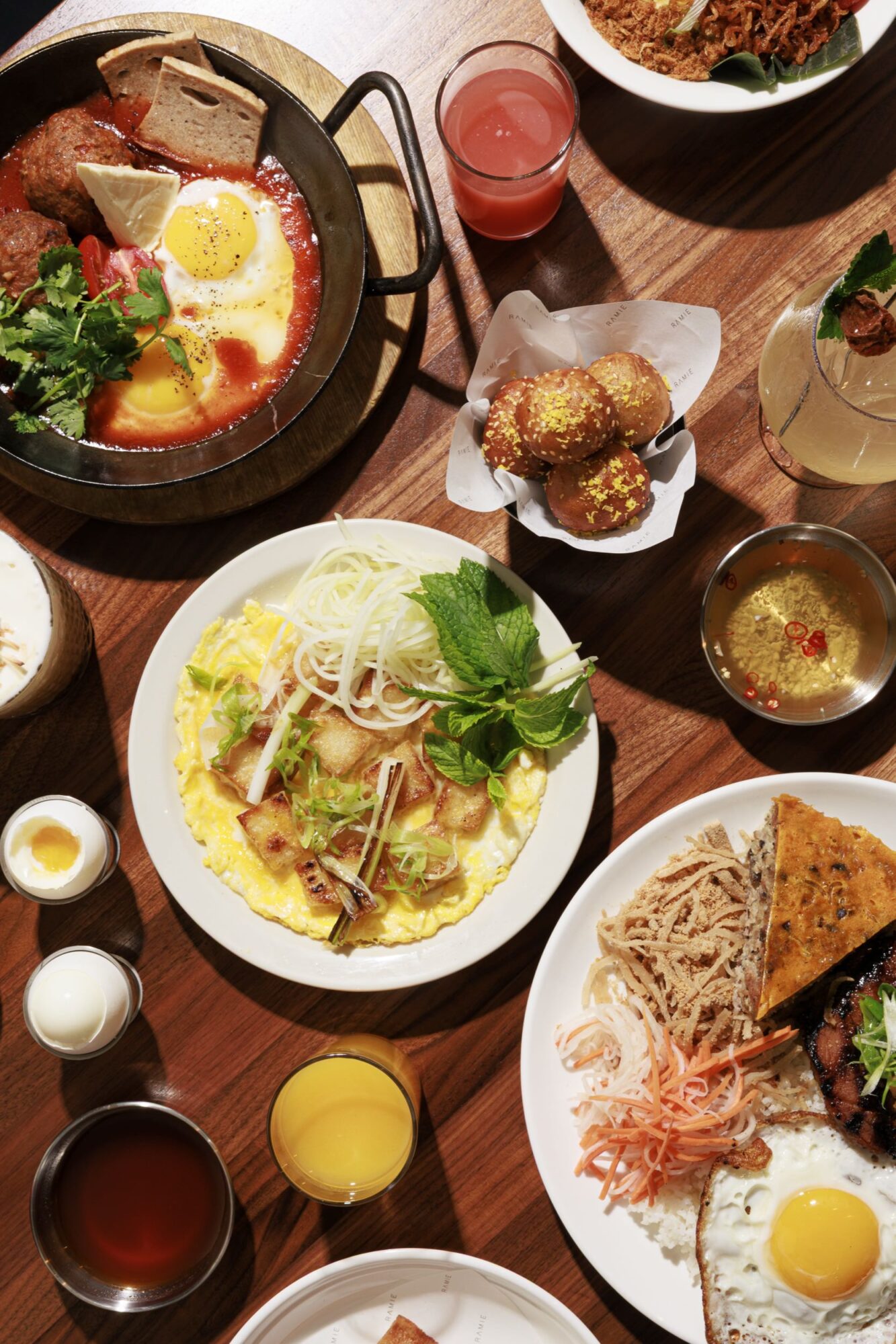 A table set with various Vietnamese dishes, including an egg skillet, omelet with herbs, crispy buns, drinks, and plates of grilled meat, noodles, and pickled vegetables. The food is colorful and attractively arranged. captured by seattle's best food and hospitality photographer Brooke Fitts