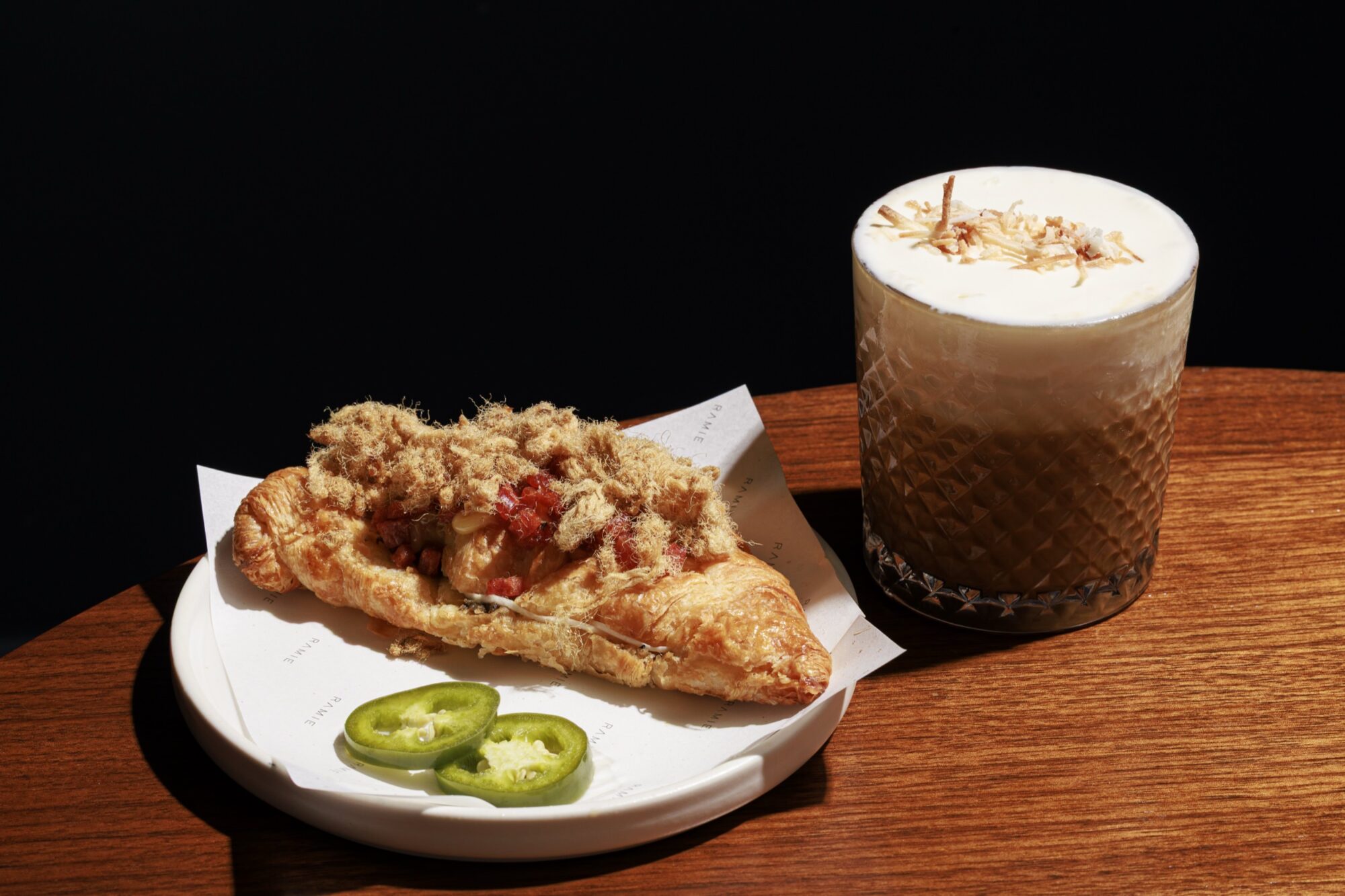 A croissant topped with meat floss, cheese, and bacon sits on a plate with sliced jalapeños, next to a glass of iced coffee with foam, on a wooden table against a dark background. captured by seattle's best food and hospitality photographer Brooke Fitts