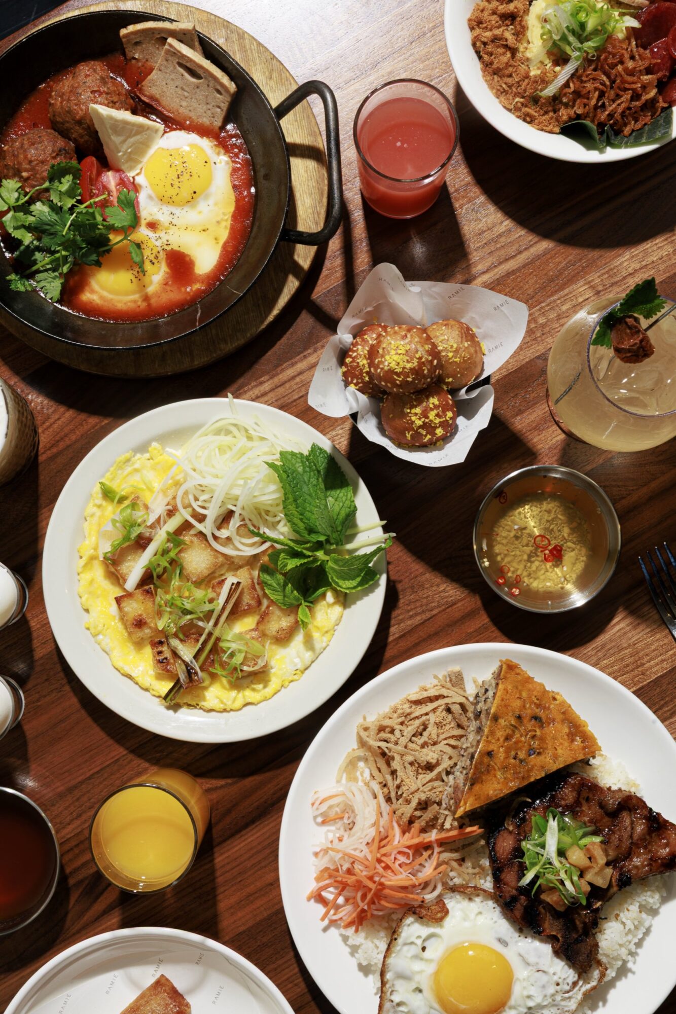 A wooden table spread with various Vietnamese dishes, including sunny-side-up eggs, grilled meat, vermicelli noodles, sauces, drinks, fresh herbs, and small round fried items, arranged in a colorful, appetizing display. captured by seattle's best food and hospitality photographer Brooke Fitts