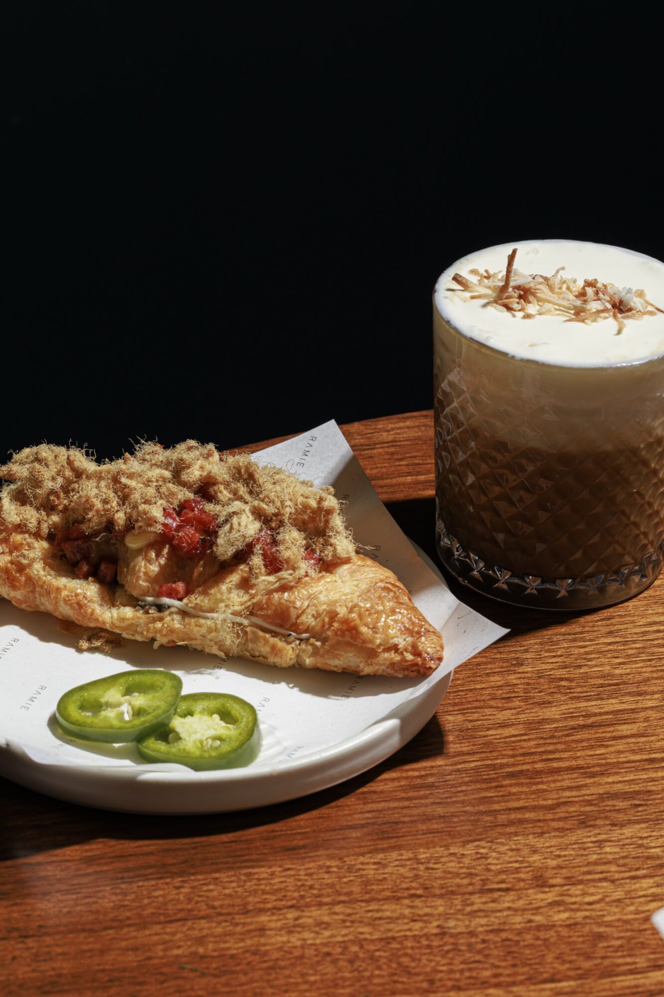 A flaky pastry topped with meat floss and diced toppings sits on a white plate beside sliced jalapeños. Next to it is a glass of iced coffee or latte, topped with foam and shredded garnish, on a wooden table. captured by seattle's best food and hospitality photographer Brooke Fitts