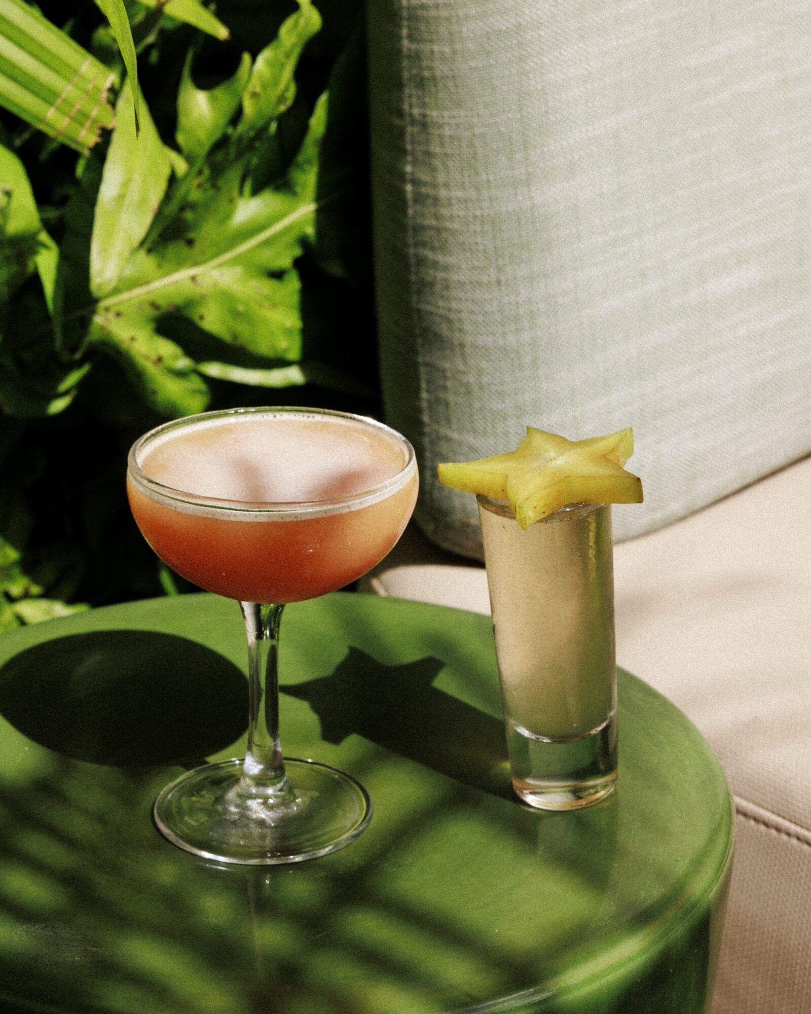 A coupe glass with a pink cocktail and a shot glass with a light drink garnished with a starfruit slice sit on a green table next to a beige cushion and leafy green plants. captured by seattle's best food and hospitality photographer Brooke Fitts