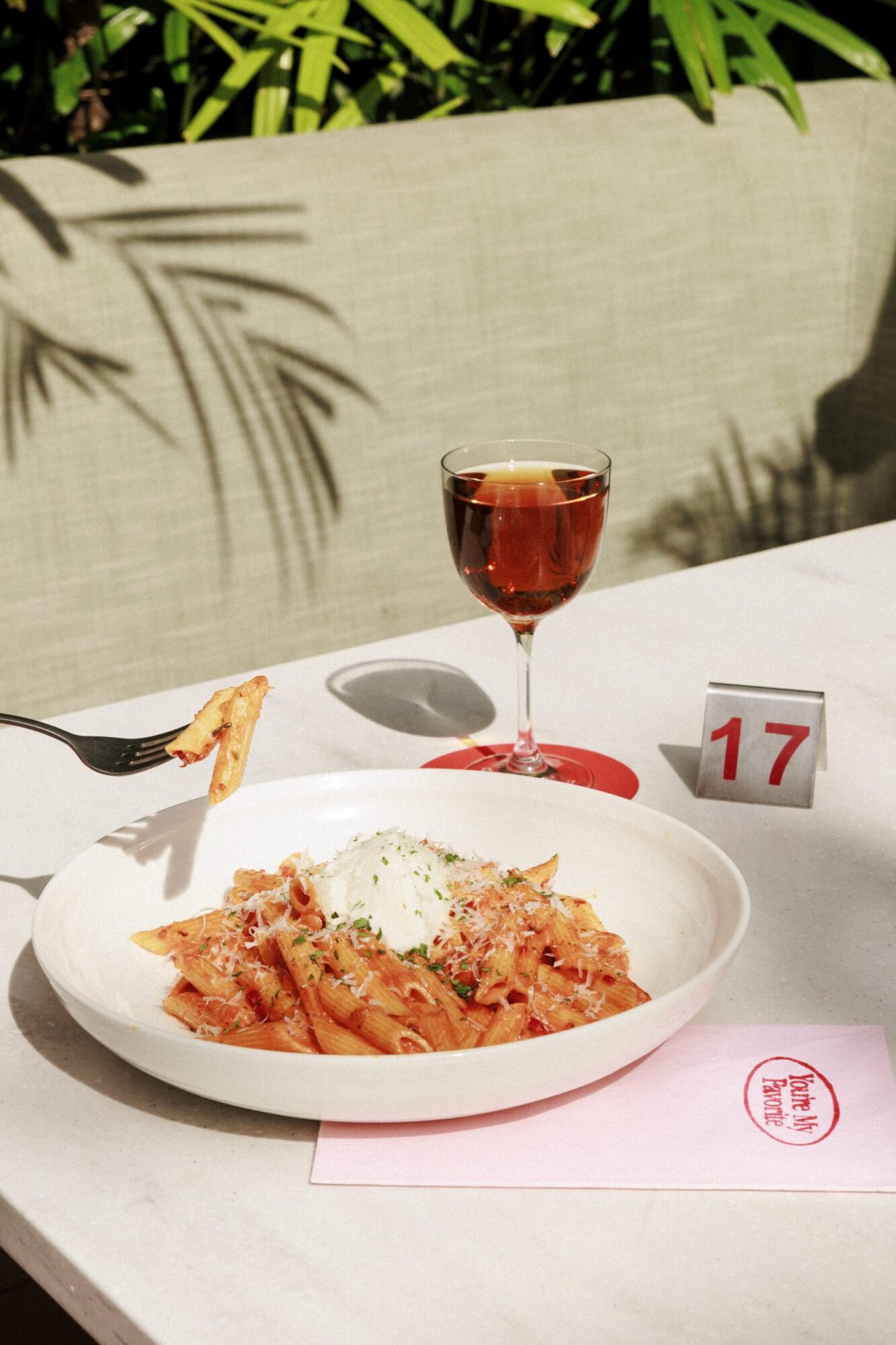 A bowl of penne pasta topped with a scoop of ricotta sits on a table beside a glass of rosé, a pink menu, and a table number 17 sign. A fork lifts a piece of pasta, and green plants are visible in the background. captured by seattle's best food and hospitality photographer Brooke Fitts