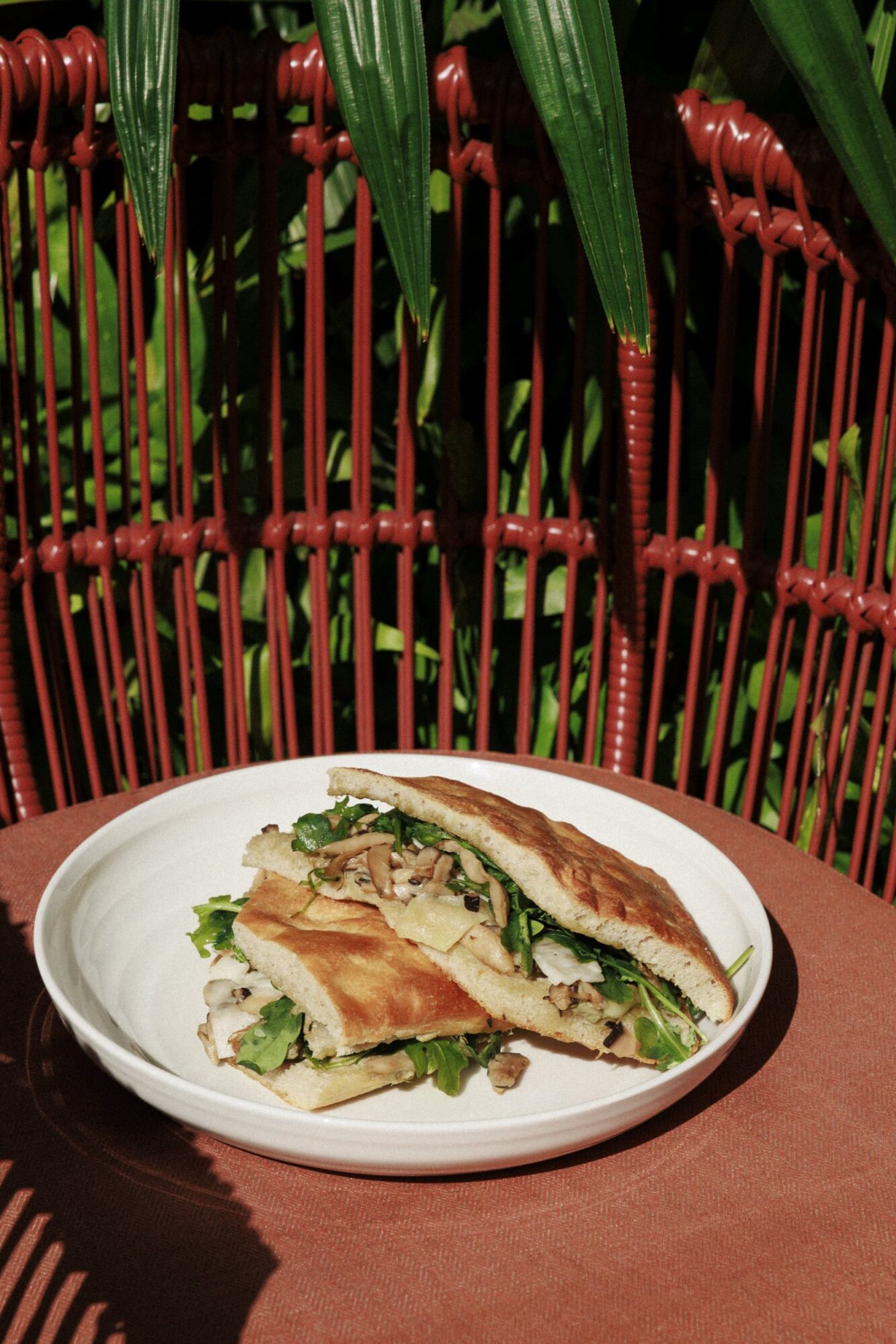 A white plate holds two halves of a sandwich filled with greens and sliced mushrooms, set on a rust-colored table with a red wicker chair and green plants in the background. captured by seattle's best food and hospitality photographer Brooke Fitts