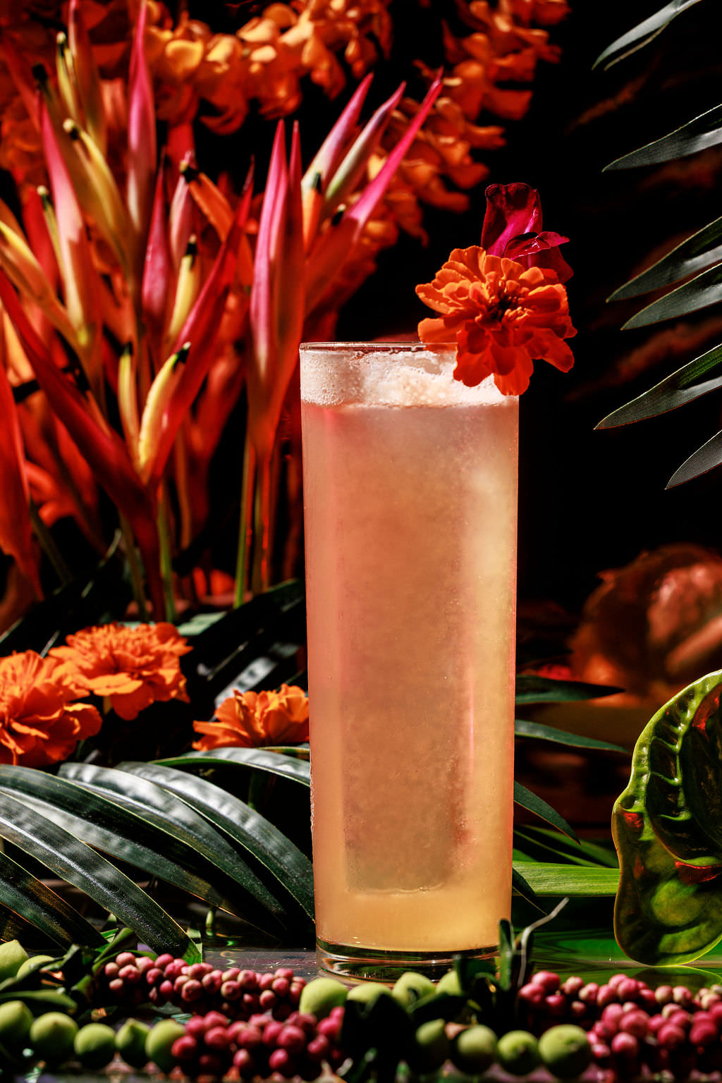 A tall glass of a light orange cocktail with ice, garnished with a vibrant orange flower, stands amid lush tropical foliage and vivid orange and red flowers. captured by seattle's best food and hospitality photographer Brooke Fitts