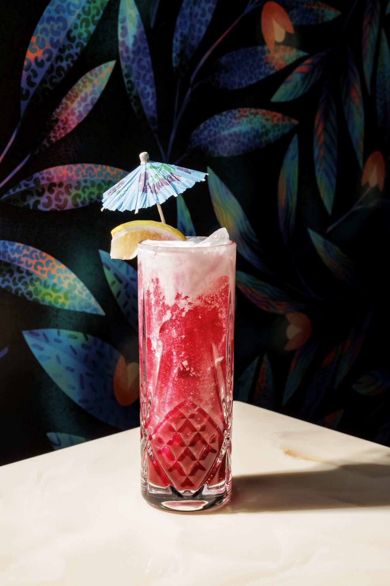 A tall glass of bright red, foamy cocktail with ice, garnished with a lemon wedge and a blue paper umbrella, sits on a light surface against a colorful, leafy patterned background. captured by seattle's best food and hospitality photographer Brooke Fitts