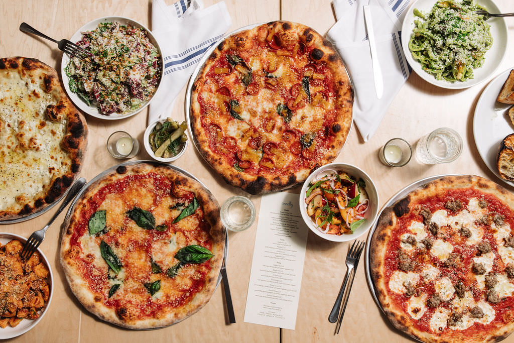 Overhead view of a table set with four pizzas, two salads, a pasta dish, bread, and drinks. A restaurant menu sits in the center, surrounded by white napkins and silverware. captured by seattle's best food and hospitality photographer Brooke Fitts