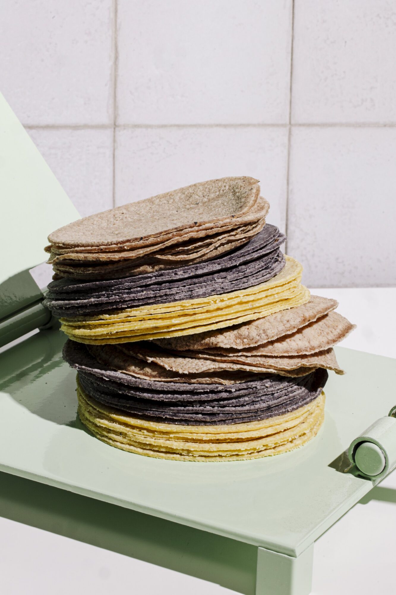 A stack of multicolored tortillas, including yellow, blue, and beige varieties, sits on a light green surface against a light tiled background. captured by seattle's best food and hospitality photographer Brooke Fitts