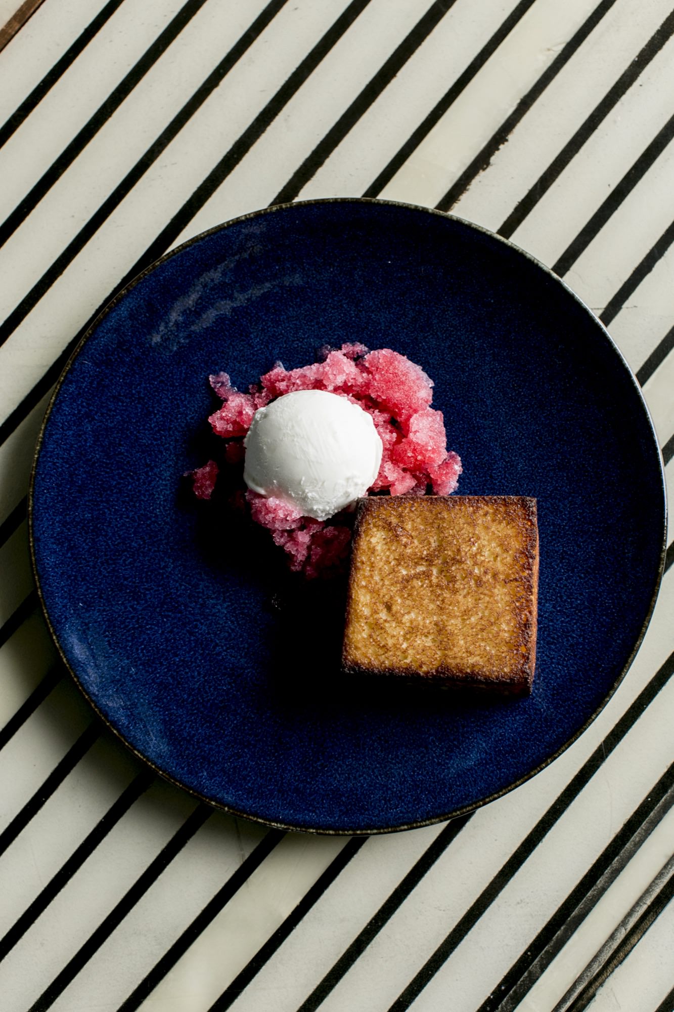 A blue plate with a brown rectangular cake, a scoop of white ice cream, and pink granita, placed on a white table with black diagonal stripes. captured by seattle's best food and hospitality photographer Brooke Fitts