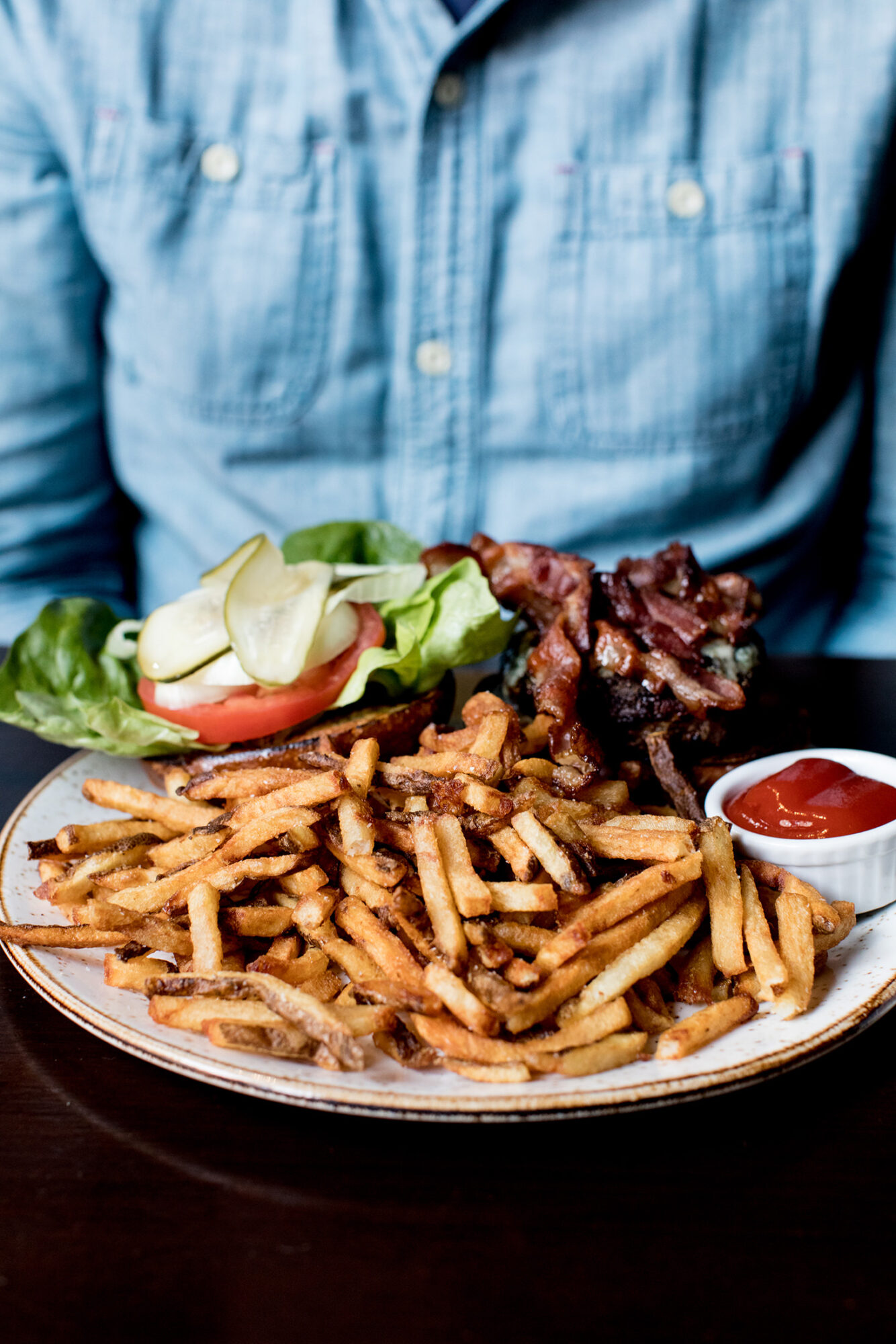A plate of French fries, a small dish of ketchup, and a lettuce, tomato, pickle, and bacon burger are in front of a person wearing a blue denim shirt. captured by seattle's best food and hospitality photographer Brooke Fitts