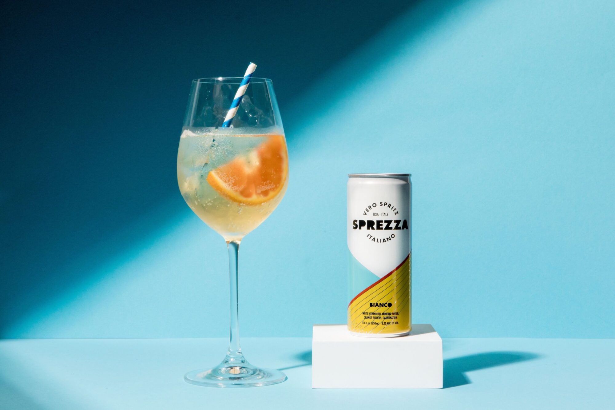 A glass of sparkling drink with ice, an orange slice, and a blue-striped straw stands next to a can of Sprezza Bianco on a blue background with bright lighting. captured by seattle's best food and hospitality photographer Brooke Fitts
