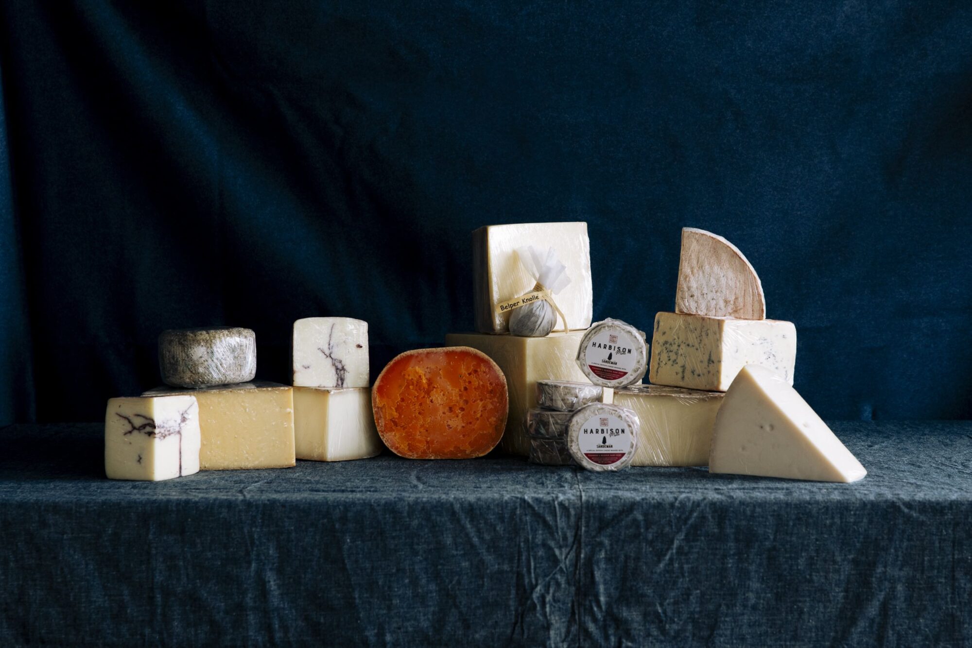 A variety of cheeses in different shapes, sizes, and colors are arranged on a blue tablecloth against a dark blue background. Some cheeses are labeled, and textures range from creamy to hard and crumbly. captured by seattle's best food and hospitality photographer Brooke Fitts