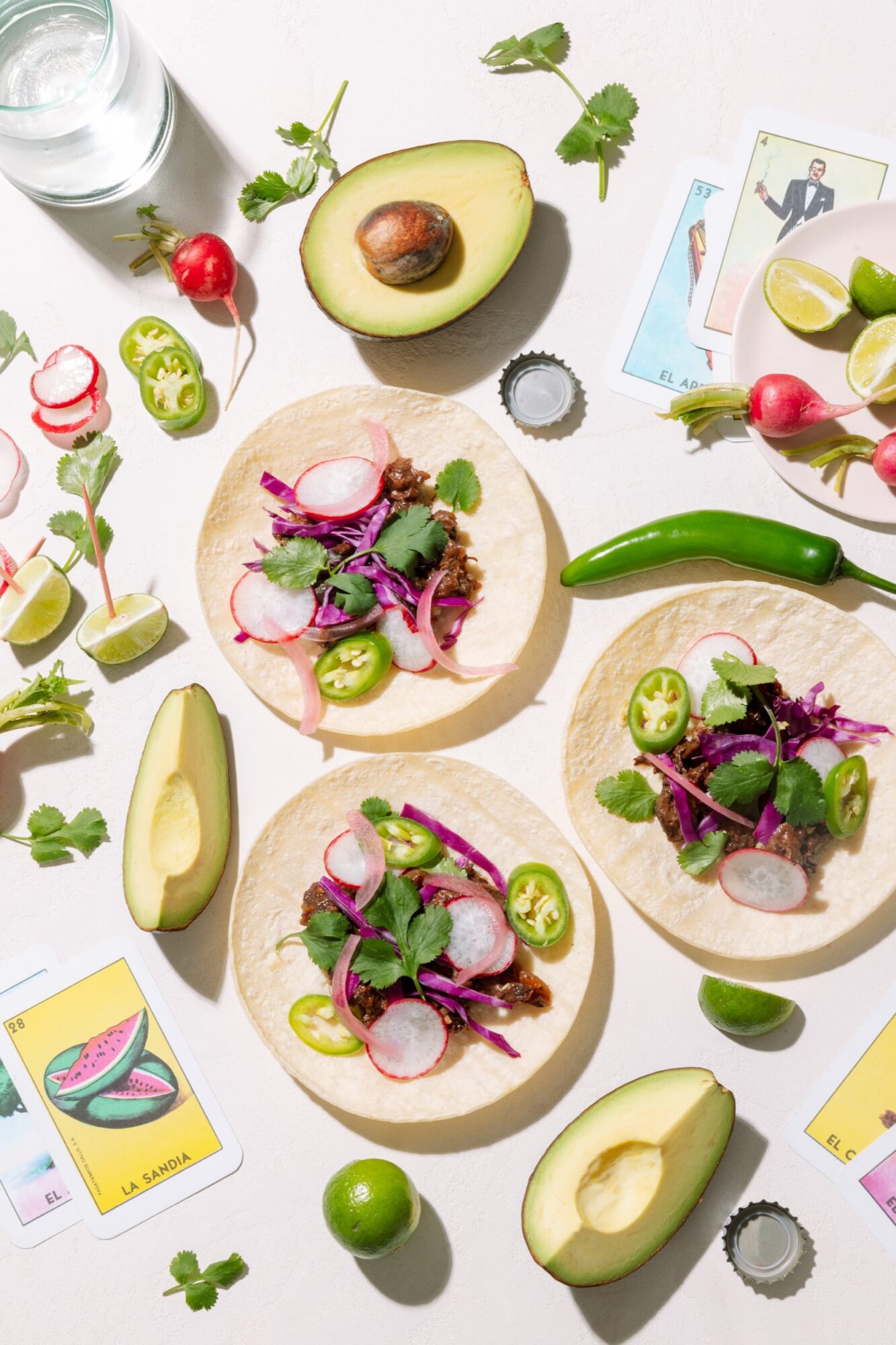 Three tacos with shredded meat, radish, cabbage, cilantro, and jalapeño on tortillas are surrounded by avocado halves, lime, radishes, jalapeños, playing cards, and a glass of water on a light surface. captured by seattle's best food and hospitality photographer Brooke Fitts