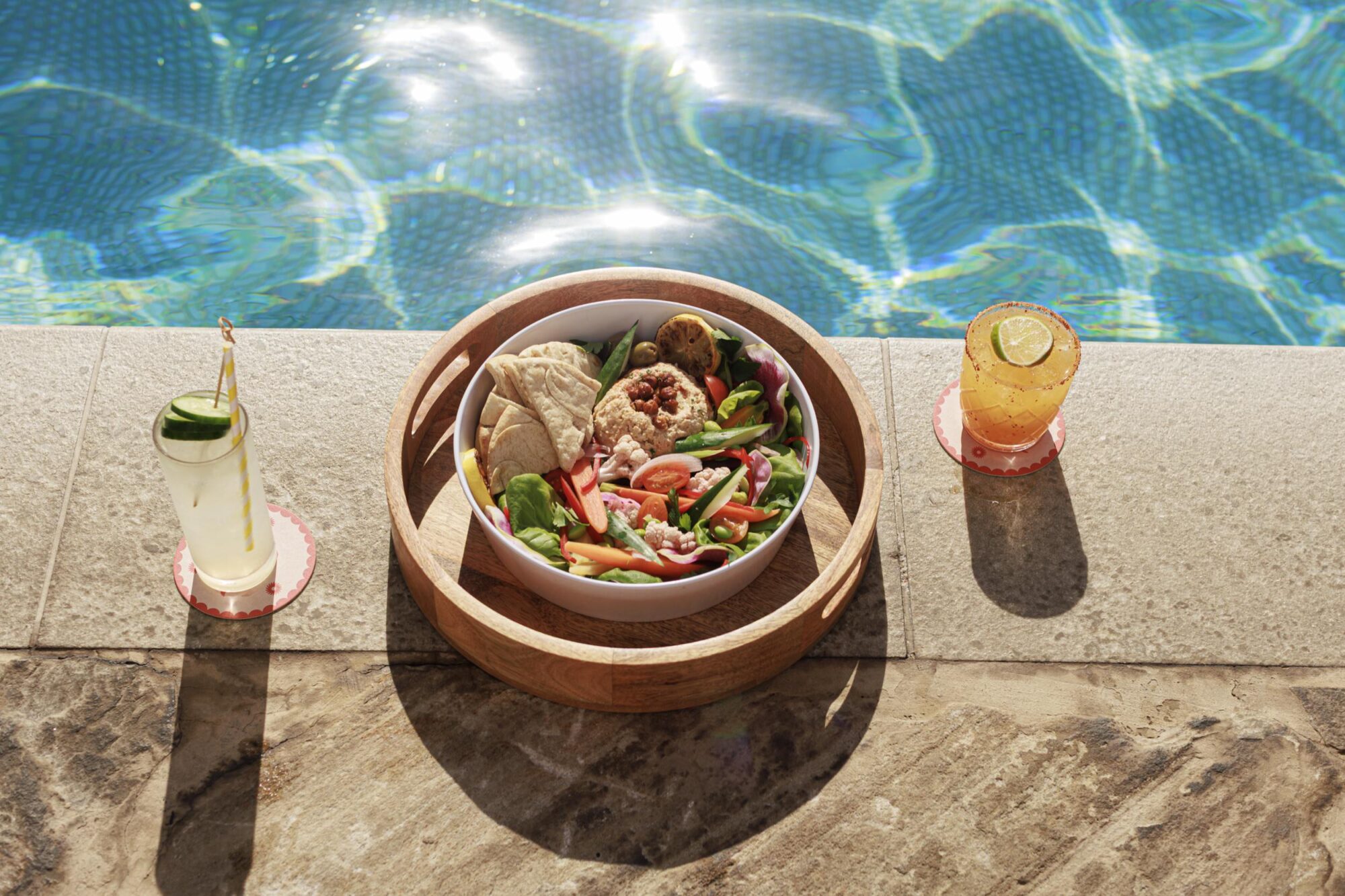 A bowl of salad with pita bread sits on a wooden tray by a pool, flanked by two cold drinks, one with a cucumber slice and straw and the other with a lemon slice. The water is bright blue and sparkling in the sunlight. captured by seattle's best food and hospitality photographer Brooke Fitts