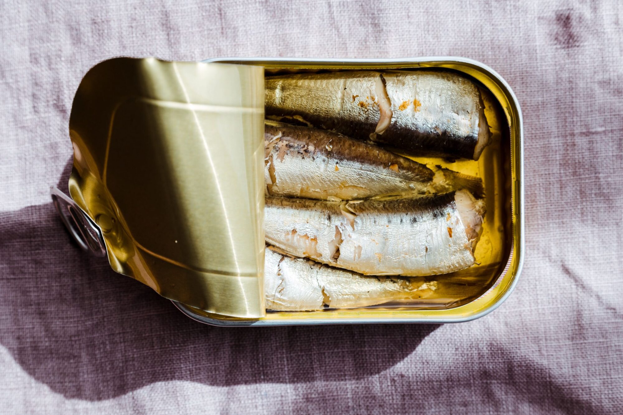 An opened tin can of sardines in oil sits on a light pink cloth, with the lid partially peeled back to reveal several whole fish packed inside. captured by seattle's best food and hospitality photographer Brooke Fitts