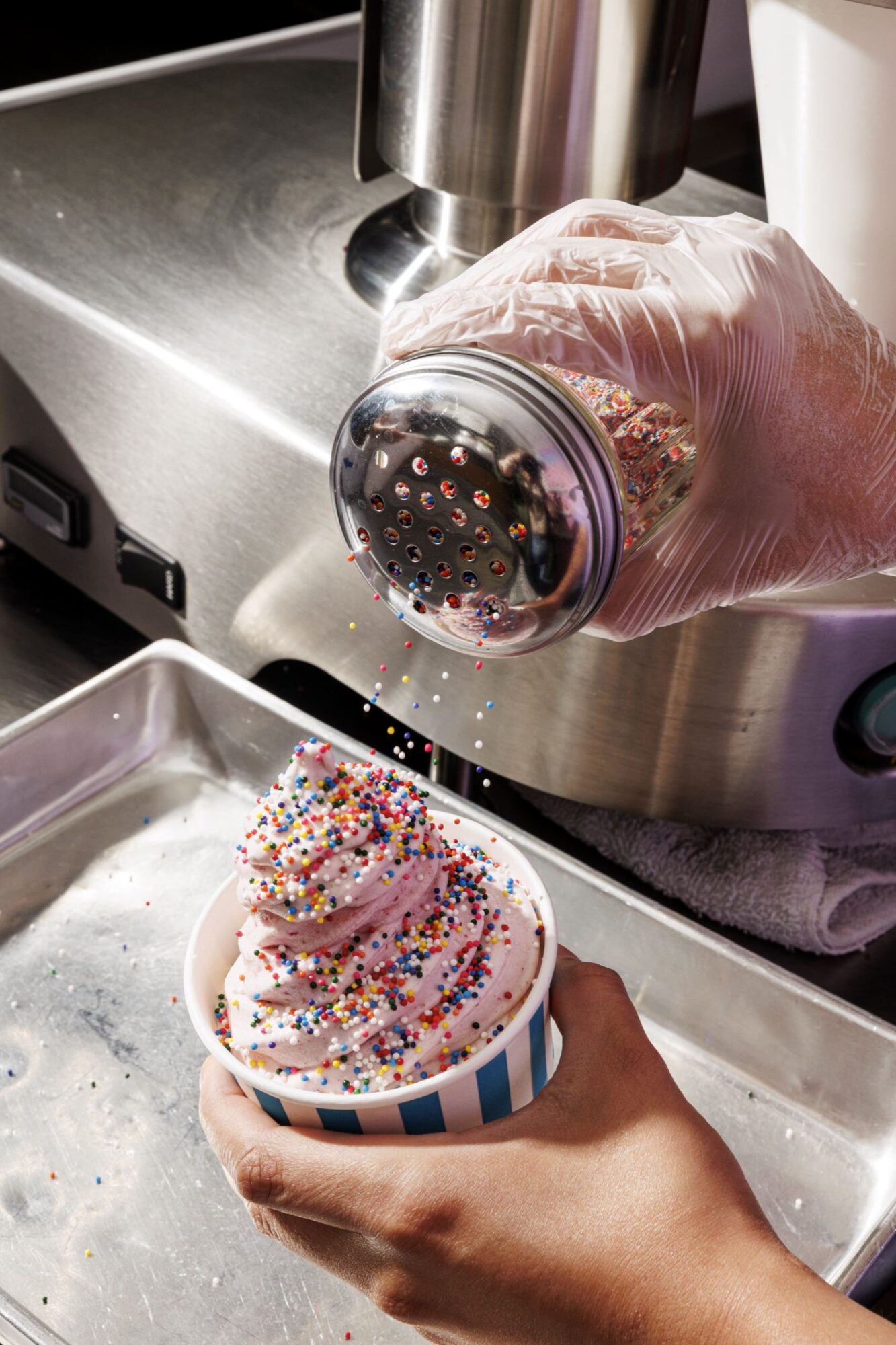 A gloved hand sprinkles colorful round sprinkles onto a cup of soft-serve ice cream, held in a blue and white striped cup, next to a metal tray and ice cream machine. captured by seattle's best food and hospitality photographer Brooke Fitts