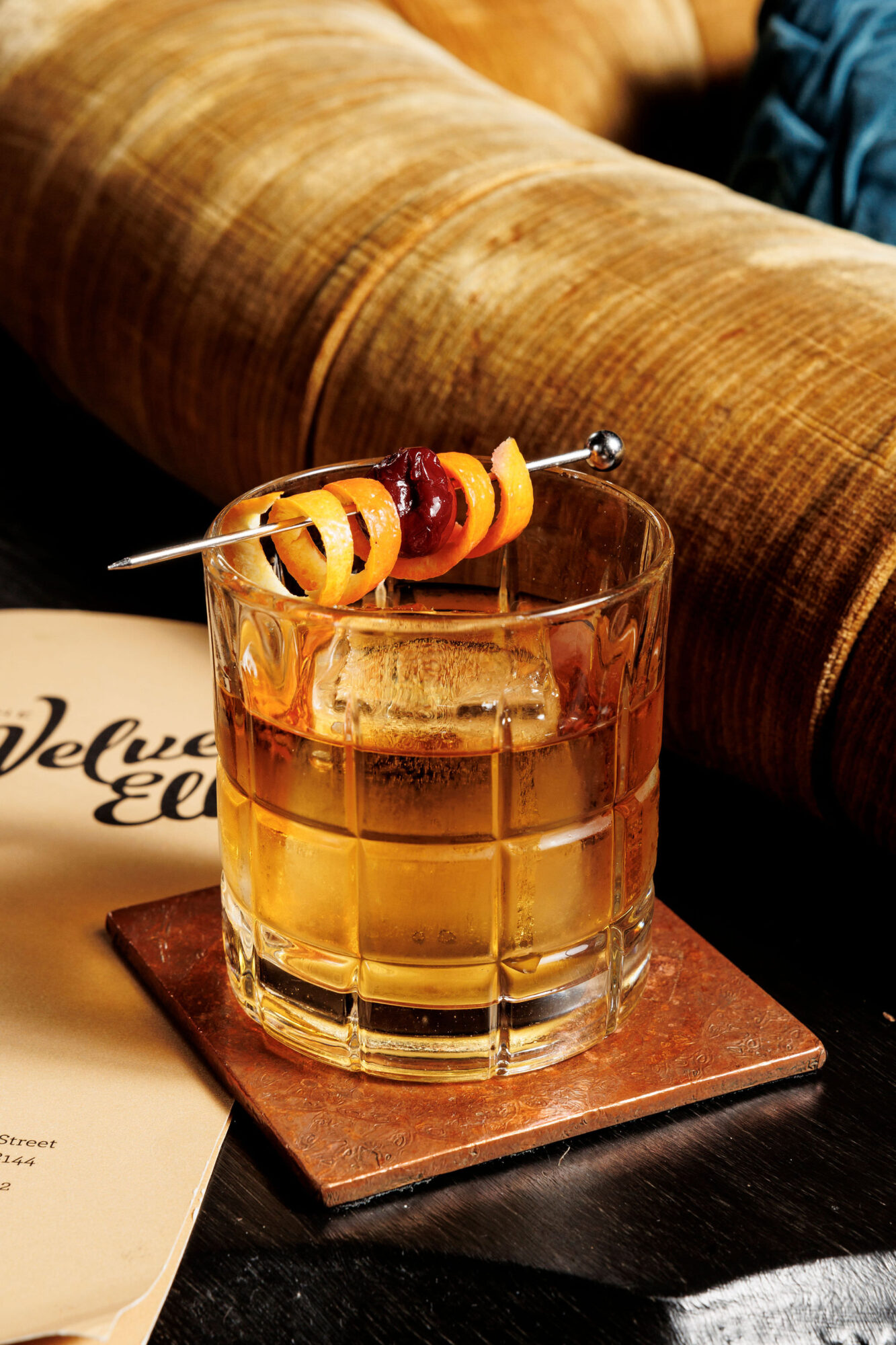 A glass of whiskey with a large ice cube, garnished with an orange twist and a cherry on a metal pick, sits on a coaster next to a menu and a plush, tan couch. captured by seattle's best food and hospitality photographer Brooke Fitts