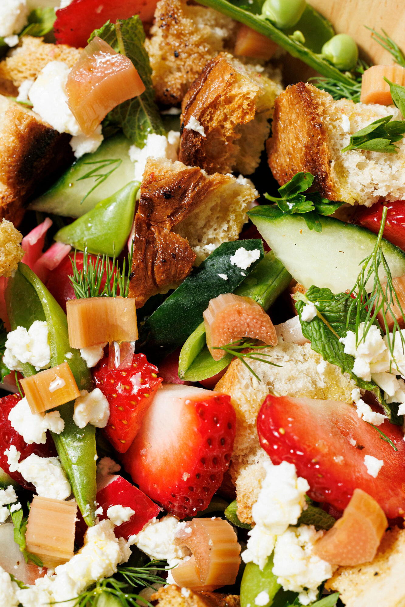 A close-up of a fresh salad with strawberries, snap peas, sliced cucumbers, crumbled feta, herbs, chunks of bread, and pieces of pink rhubarb. captured by seattle's best food and hospitality photographer Brooke Fitts