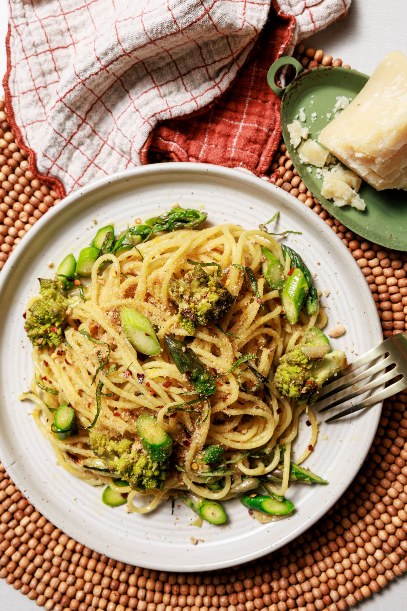 A plate of spaghetti with green vegetables, topped with grated cheese and herbs, sits on a woven placemat. A fork rests on the plate. In the background, a wedge of cheese and kitchen towels are visible. captured by seattle's best food and hospitality photographer Brooke Fitts