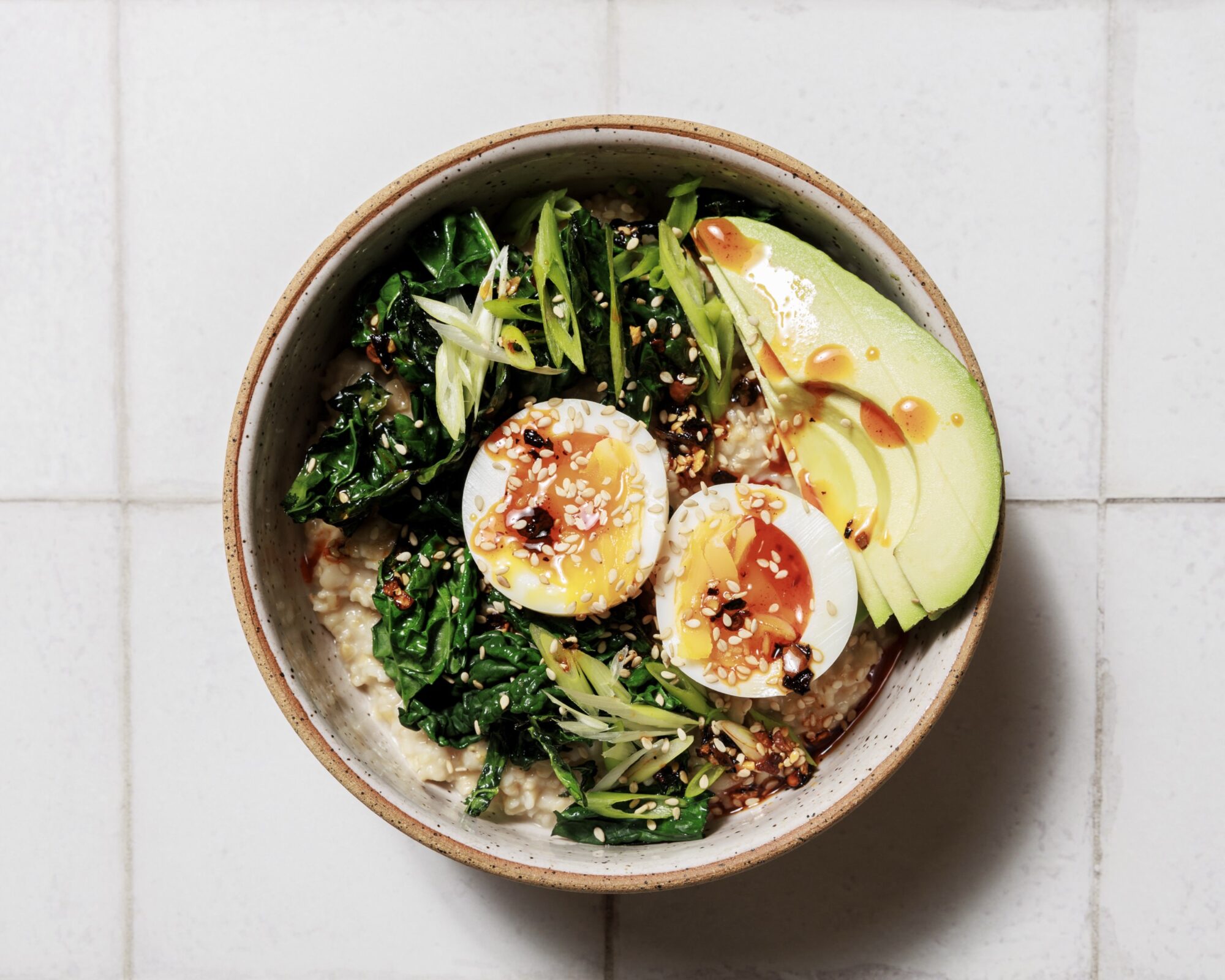 A ceramic bowl with oatmeal, topped with sliced avocado, halved soft-boiled egg, sautéed greens, chopped scallions, sesame seeds, and a drizzle of sauce, on a white tiled background. captured by seattle's best food and hospitality photographer Brooke Fitts