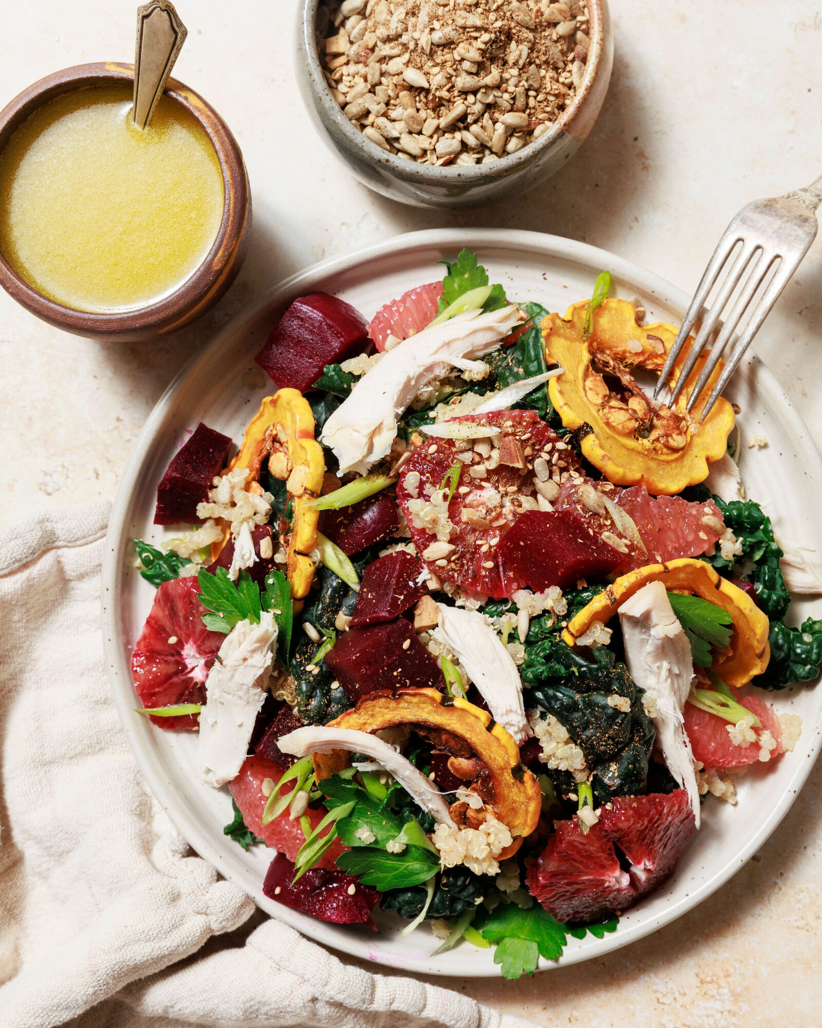 A plate of salad with shredded chicken, roasted squash, beets, citrus slices, greens, and grains, garnished with seeds. Nearby are a bowl of dressing with a spoon and a bowl of sunflower seeds. captured by seattle's best food and hospitality photographer Brooke Fitts