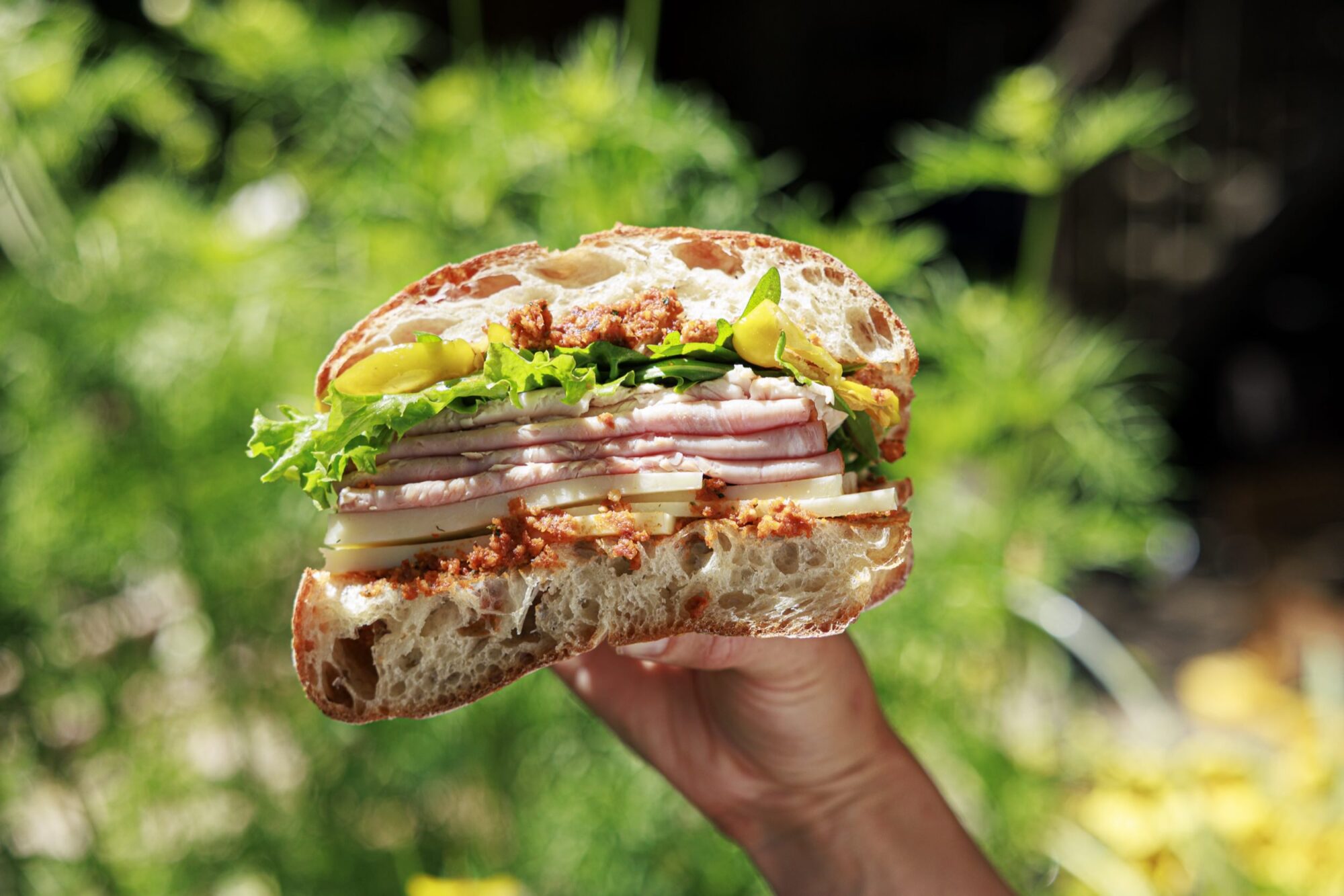 A hand holds up a thick sandwich filled with layers of ham, cheese, lettuce, yellow peppers, and a red spread, against a vibrant, blurred green outdoor background. captured by seattle's best food and hospitality photographer Brooke Fitts