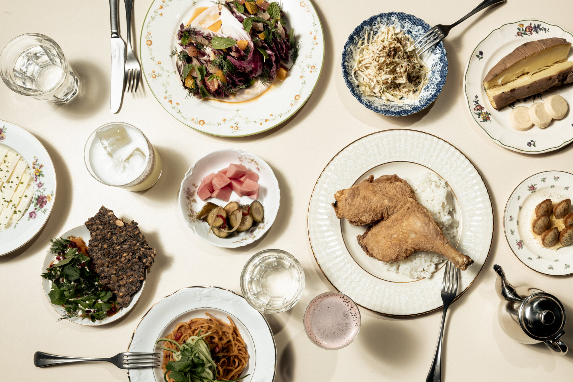 An overhead view of a table set with various dishes, including fried chicken on rice, pasta, salad, pickled vegetables, cake, bread, drinks, and other assorted plates on decorative china. captured by seattle's best food and hospitality photographer Brooke Fitts