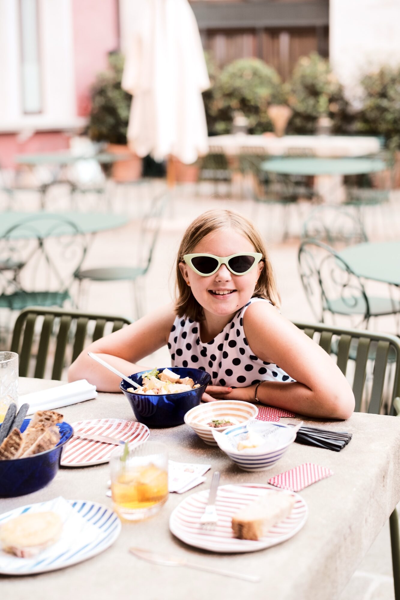 A young girl wearing white sunglasses and a polka-dot dress smiles while sitting at an outdoor café table set with bowls, plates, and drinks. The background features empty green chairs and blurred plants. captured by seattle's best food and hospitality photographer Brooke Fitts