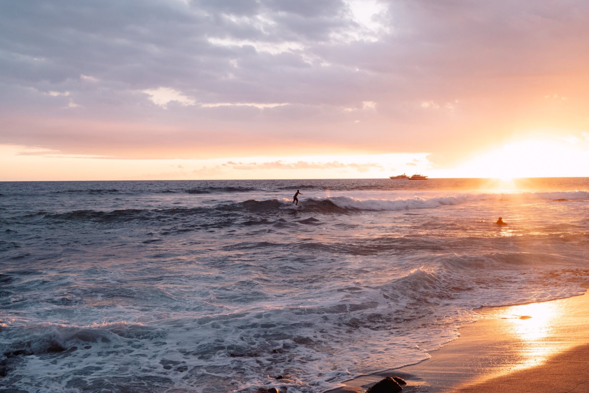 Surfers ride gentle waves near the shore at sunset, with golden sunlight reflecting on the ocean. A boat is visible on the horizon under a partly cloudy sky. captured by seattle's best food and hospitality photographer Brooke Fitts