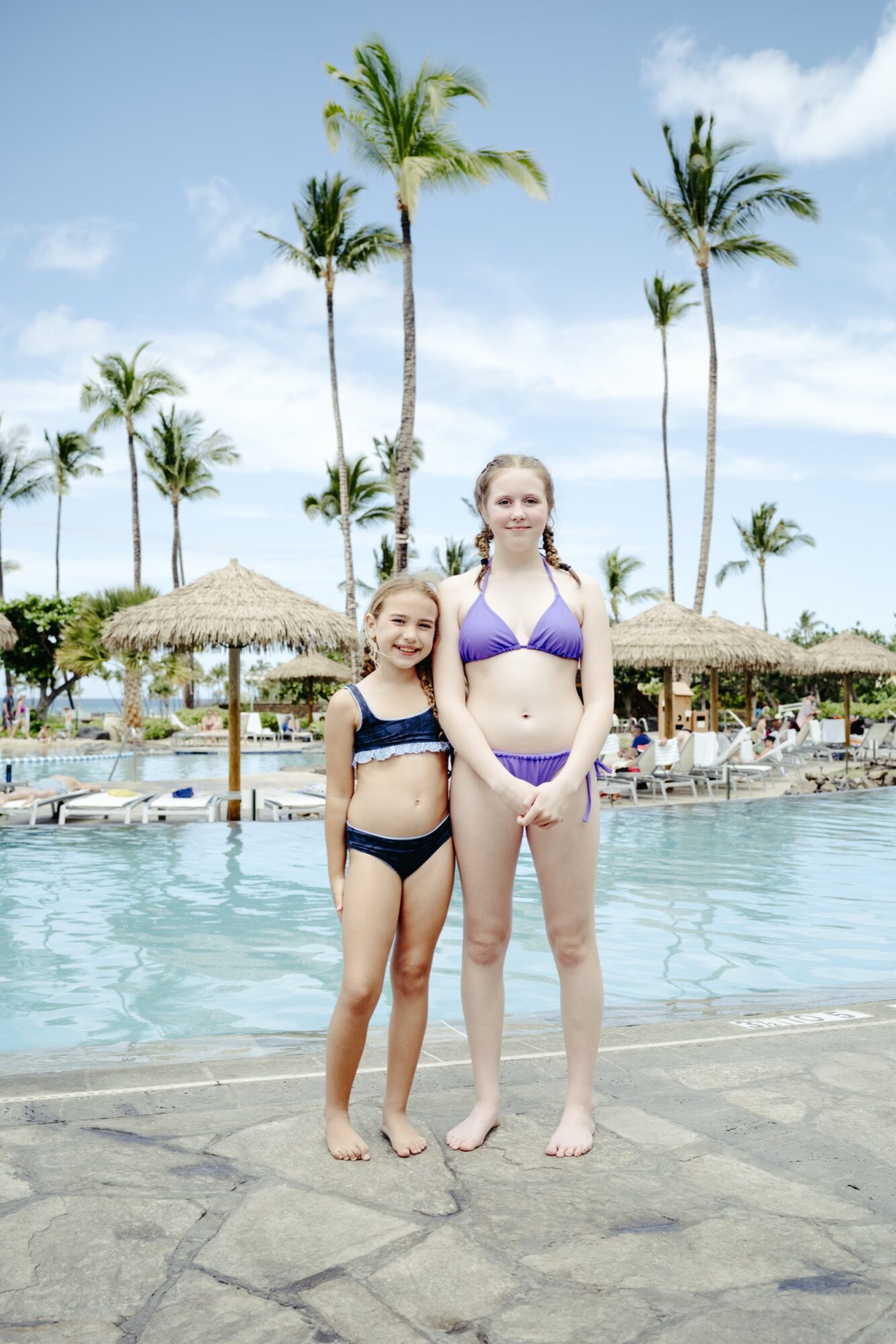 Two girls in swimsuits stand side by side by a pool with palm trees and thatched umbrellas in the background on a sunny day. The setting appears tropical and relaxed, with lounge chairs and blue sky visible. captured by seattle's best food and hospitality photographer Brooke Fitts