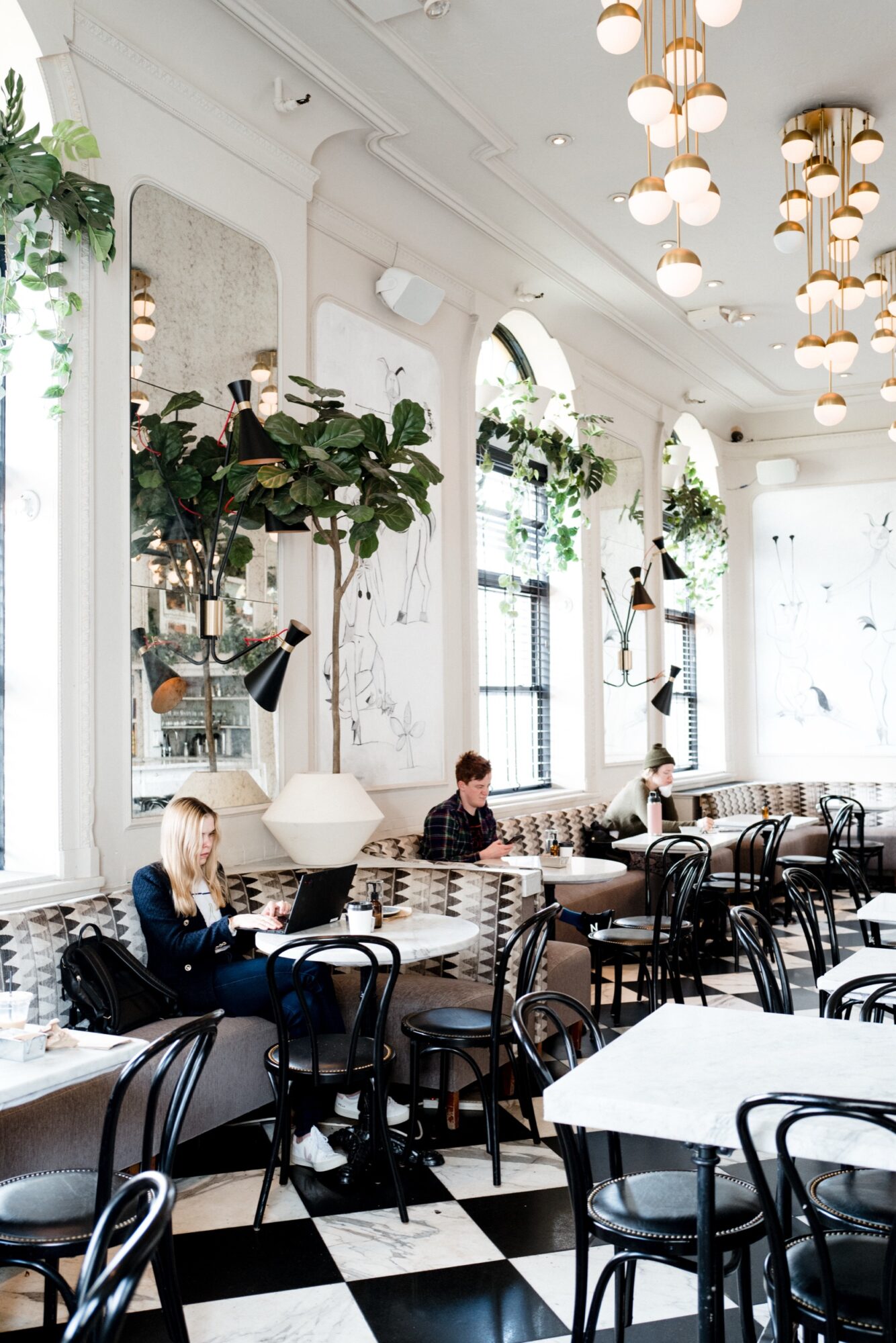 A bright, stylish café with black-and-white checkered floors, large mirrors, hanging globe lights, and several people working or relaxing at tables with laptops and drinks. captured by seattle's best food and hospitality photographer Brooke Fitts
