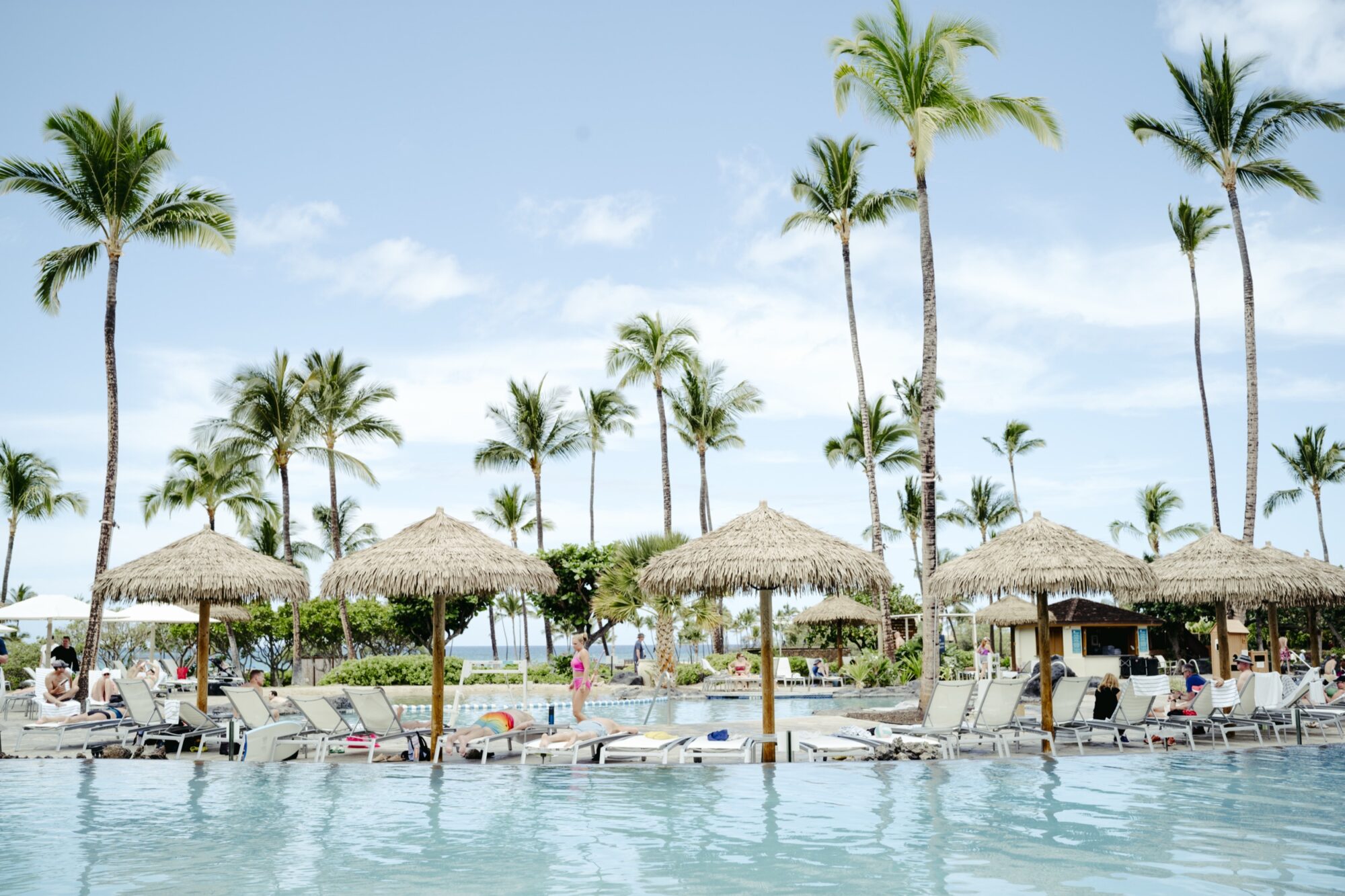 A tropical resort pool with lounge chairs, straw umbrellas, and palm trees under a blue sky. People relax by the pool, and the ocean is visible in the background. captured by seattle's best food and hospitality photographer Brooke Fitts