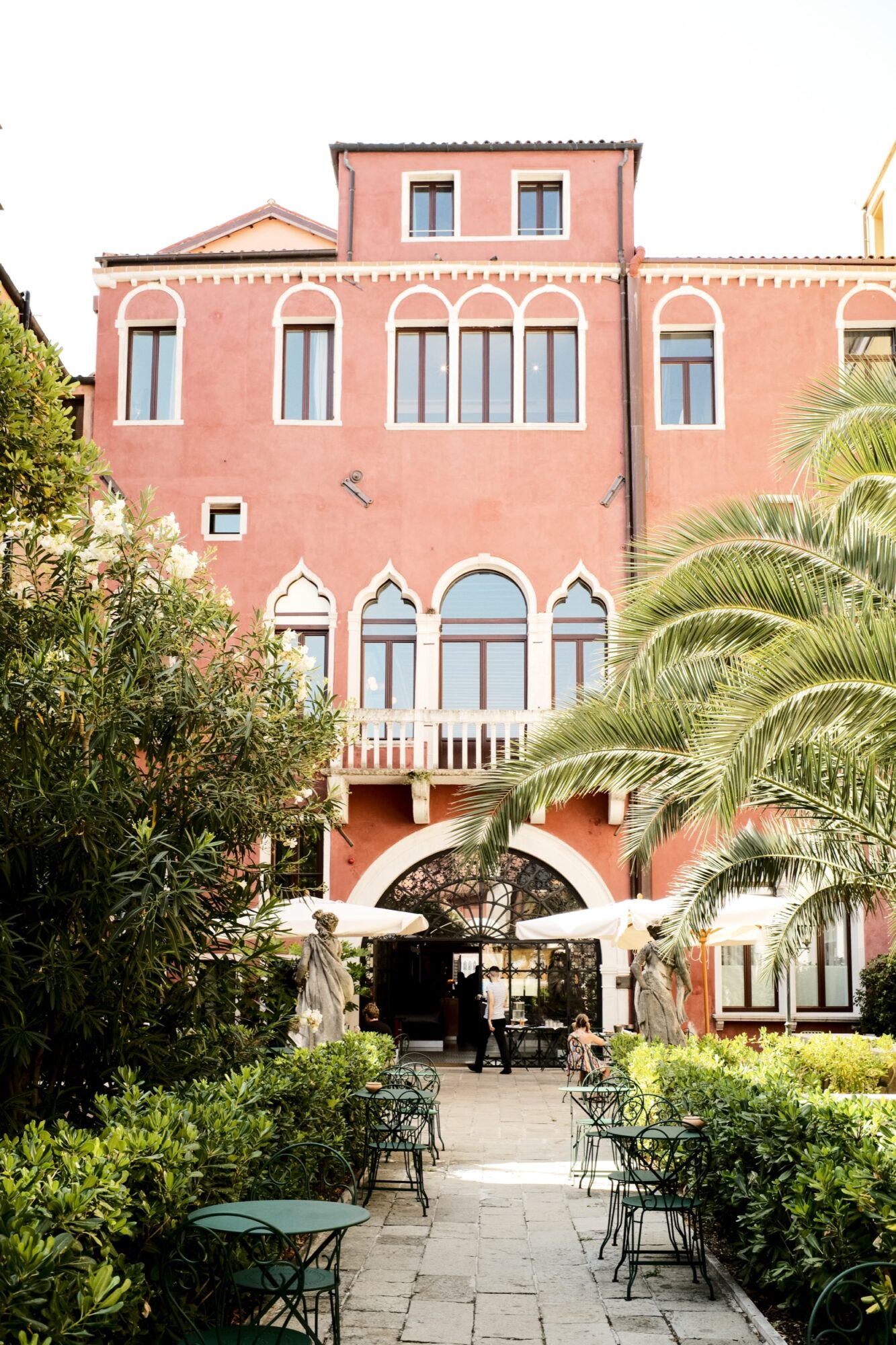 A vibrant red three-story building with arched windows and white trim; in front, a garden patio with green metal tables and chairs, surrounded by lush greenery and palm trees. captured by seattle's best food and hospitality photographer Brooke Fitts