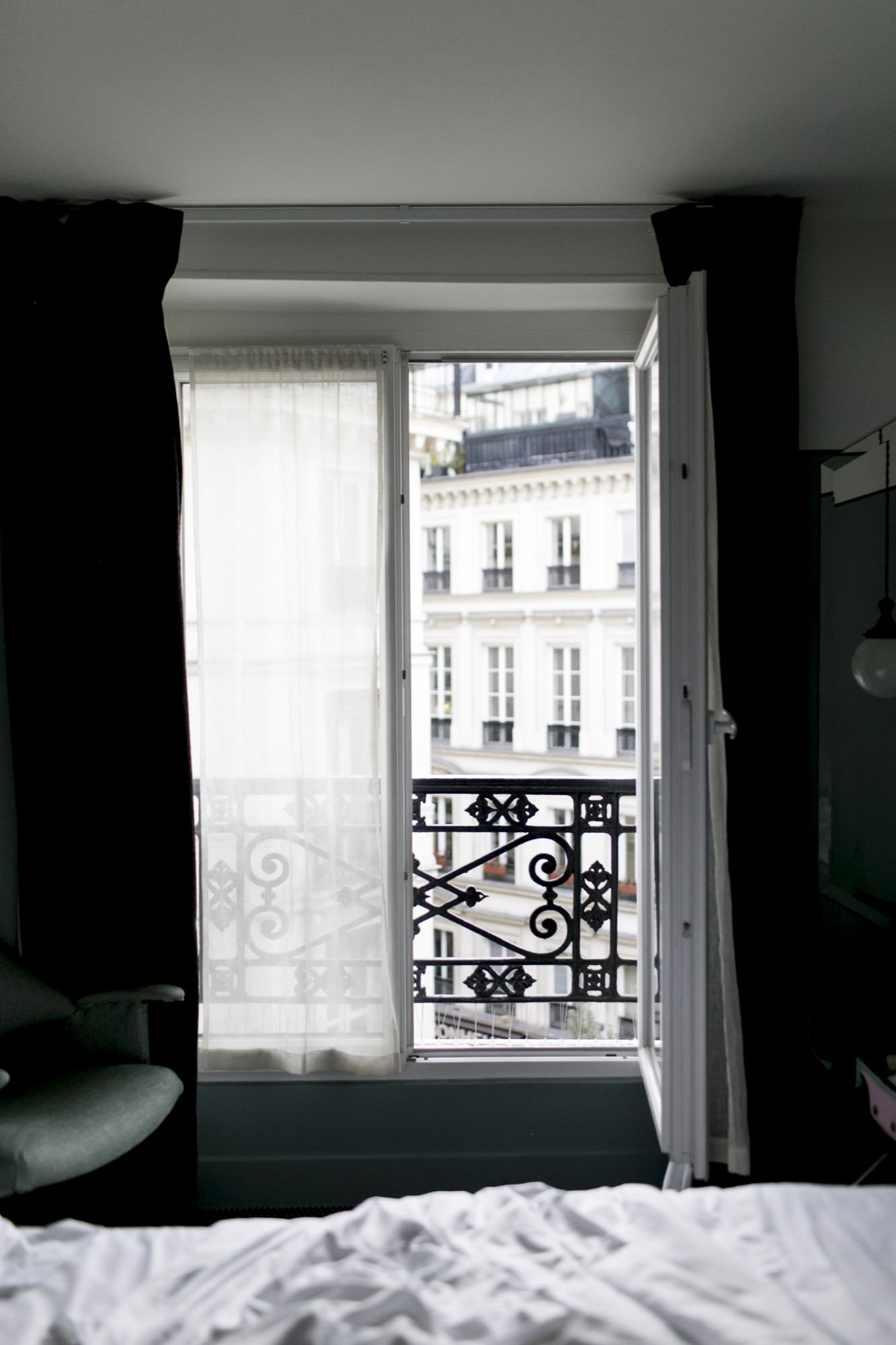 A window with sheer curtains and an ornate black railing opens to a view of white Parisian-style buildings, seen from inside a dimly lit room with a chair and part of an unmade bed. captured by seattle's best food and hospitality photographer Brooke Fitts
