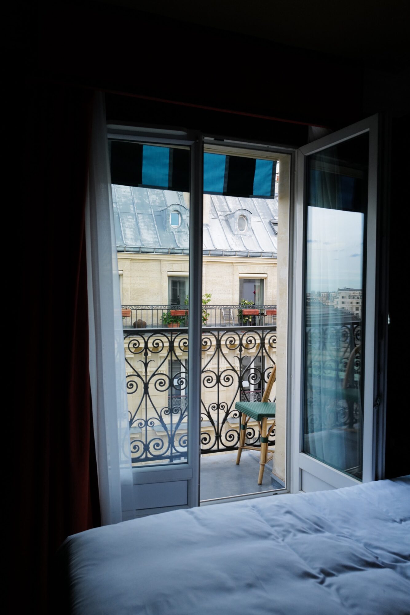 An open glass door leads to a small balcony with a decorative iron railing, a wooden chair, and potted plants, overlooking a building with a tin roof and windows. Part of an unmade bed is visible in the foreground. captured by seattle's best food and hospitality photographer Brooke Fitts