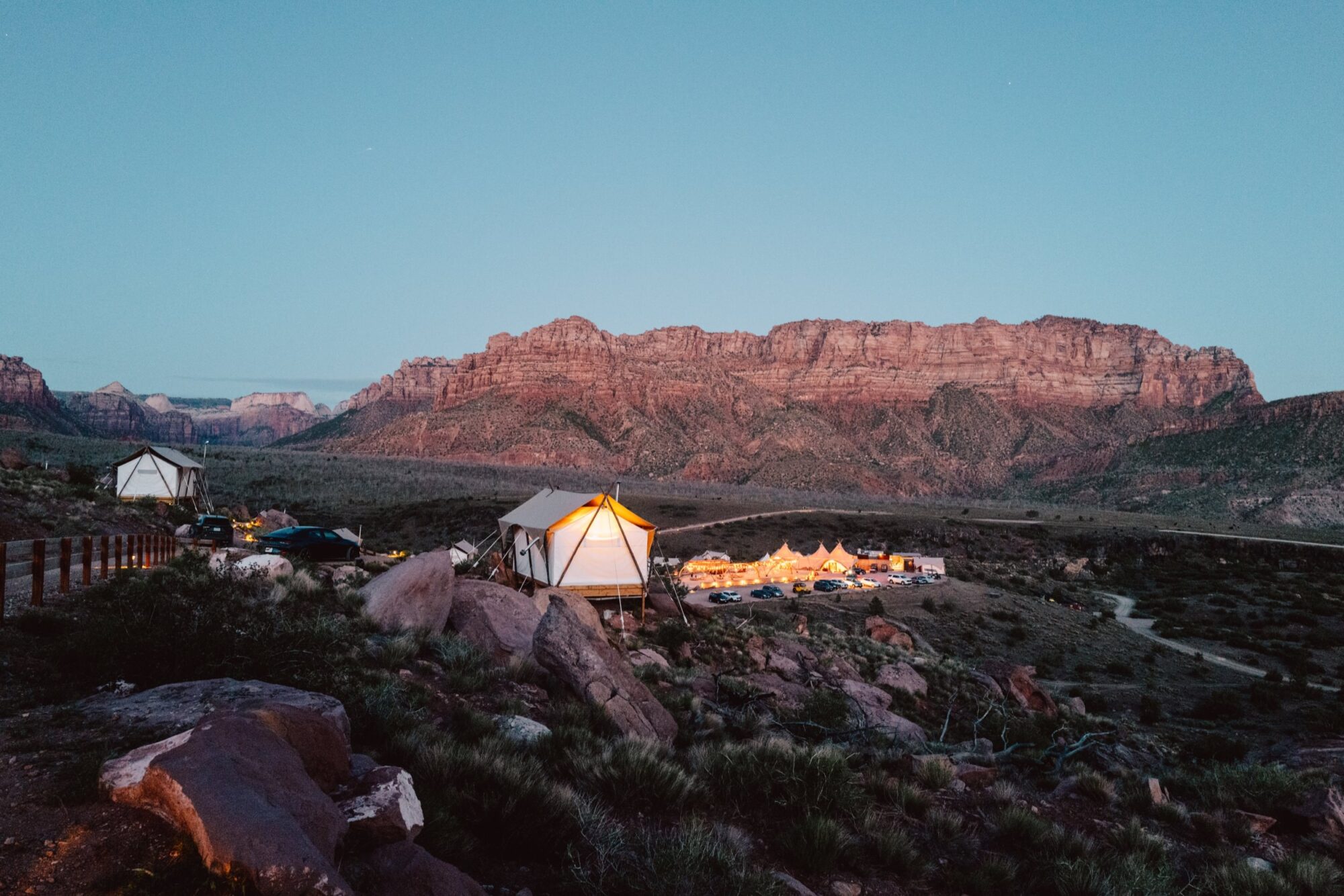 Tents and small buildings are illuminated at dusk in a rocky desert landscape, with rugged red mountains in the background and clear blue sky overhead. captured by seattle's best food and hospitality photographer Brooke Fitts