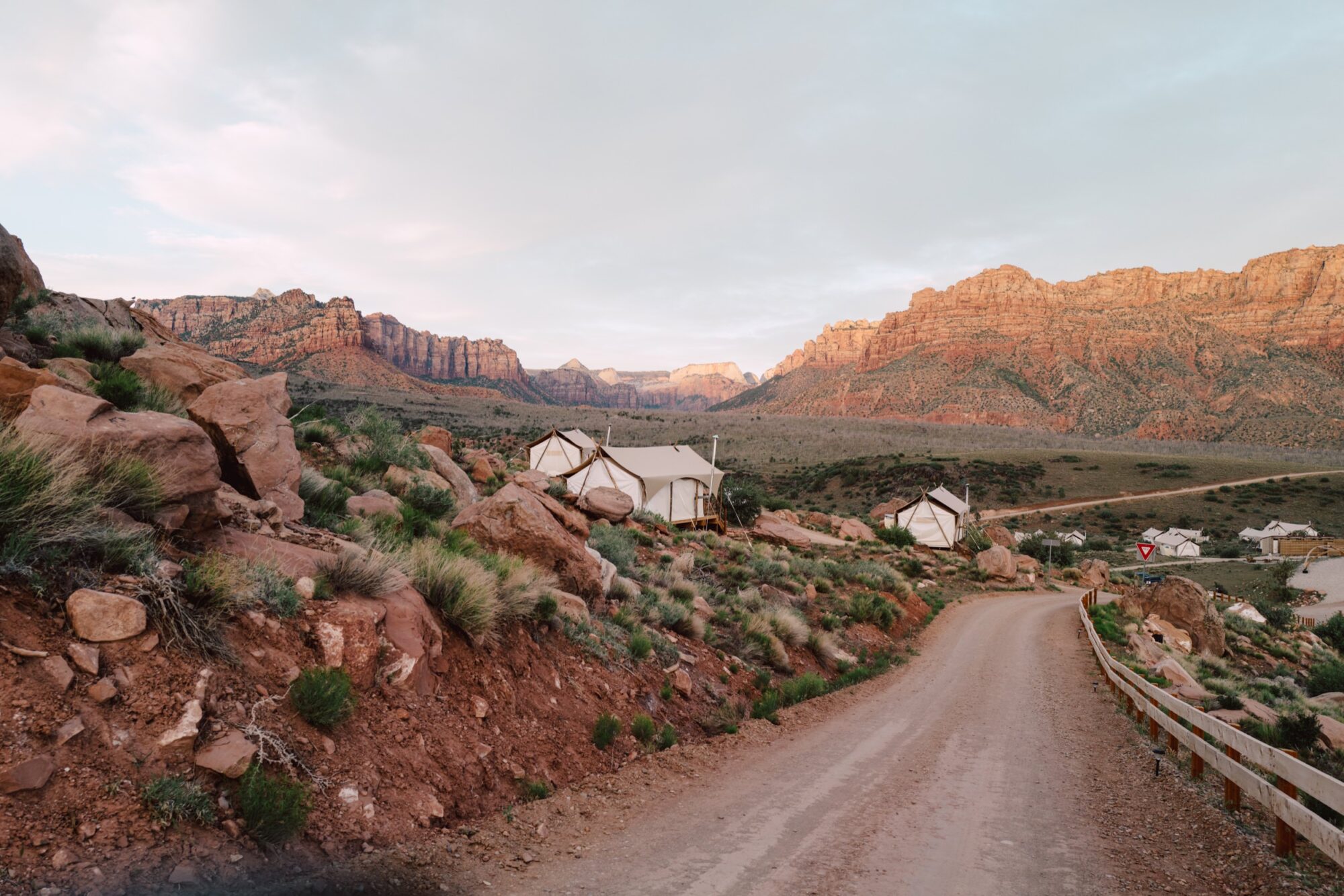 A dirt road curves past canvas tents set up on a rocky hillside with red rock mountains and desert vegetation in the background under a cloudy sky at sunset. captured by seattle's best food and hospitality photographer Brooke Fitts