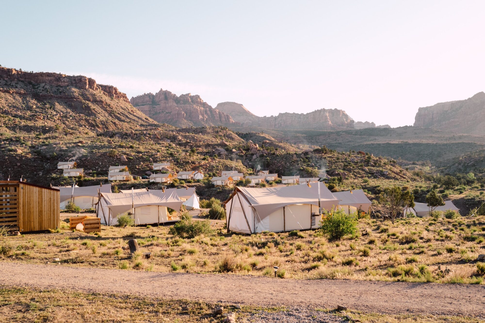 A scenic glamping site with white canvas tents spread across a grassy, desert landscape surrounded by rocky hills and mountains under a clear sky. captured by seattle's best food and hospitality photographer Brooke Fitts