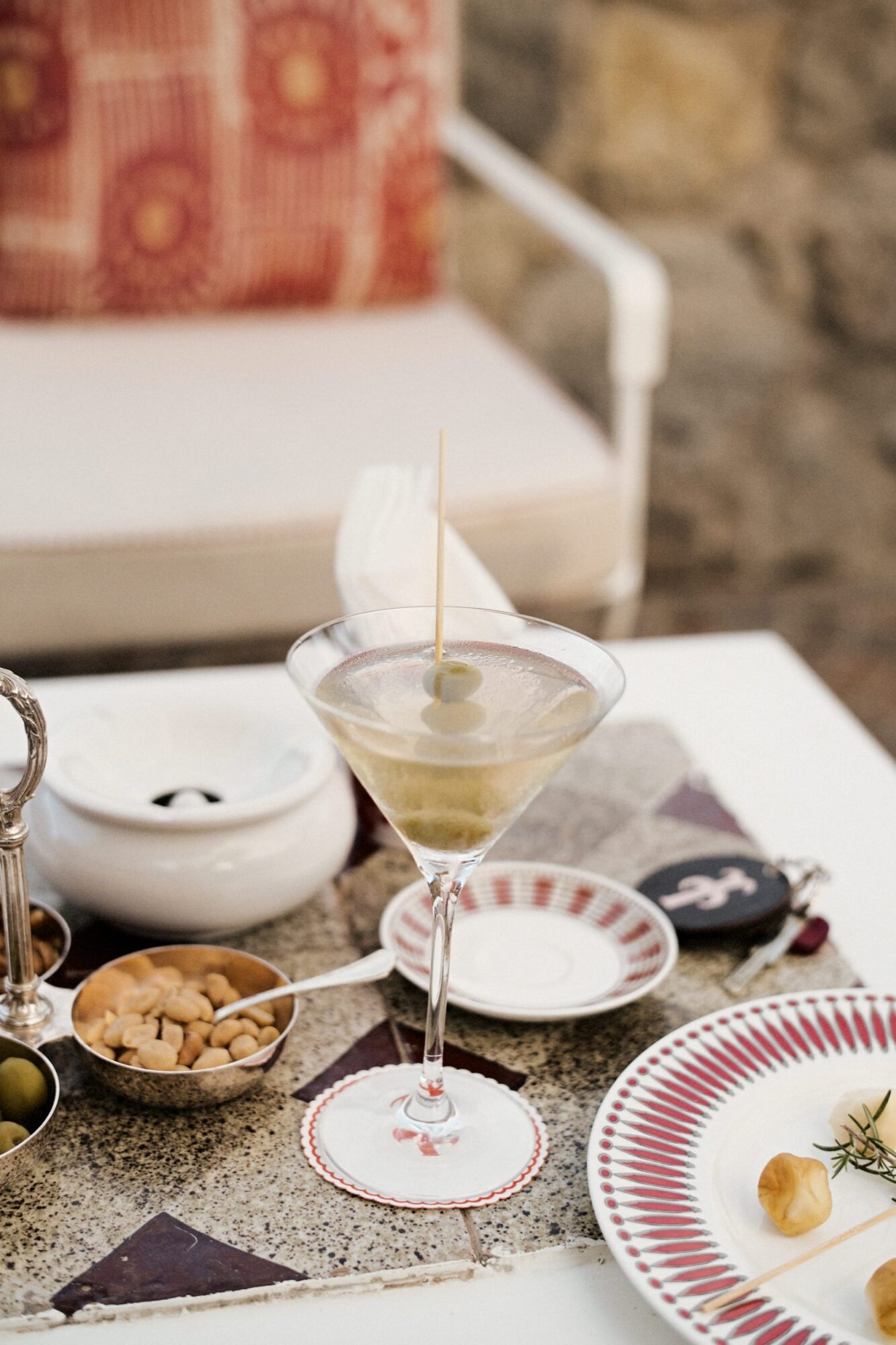 A martini with an olive garnish on a toothpick sits on a white table, surrounded by small bowls of snacks and a white ashtray. A patterned red cushion and stone wall are in the background. captured by seattle's best food and hospitality photographer Brooke Fitts