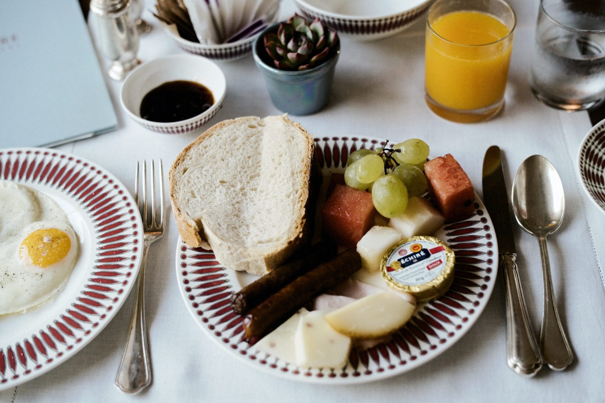A breakfast plate with bread slices, two sausages, various cheeses, grapes, melon cubes, butter, and jam. Nearby are a glass of orange juice, a cup, a plate with a fried egg, utensils, and a small potted plant. captured by seattle's best food and hospitality photographer Brooke Fitts