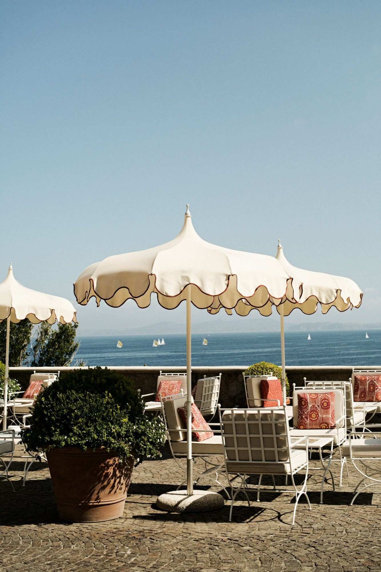 White patio umbrellas and cushioned chairs sit on a stone terrace with potted plants, overlooking a calm blue sea dotted with sailboats under a clear sky. captured by seattle's best food and hospitality photographer Brooke Fitts