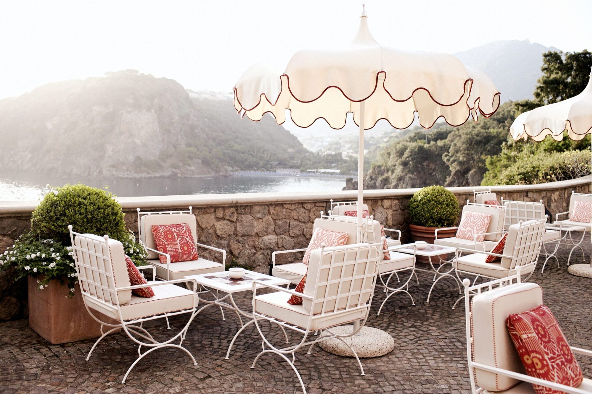 Outdoor café terrace with white metal chairs and tables, red and white cushions, large white umbrellas, potted green plants, and a scenic view of water and hills in the background. captured by seattle's best food and hospitality photographer Brooke Fitts