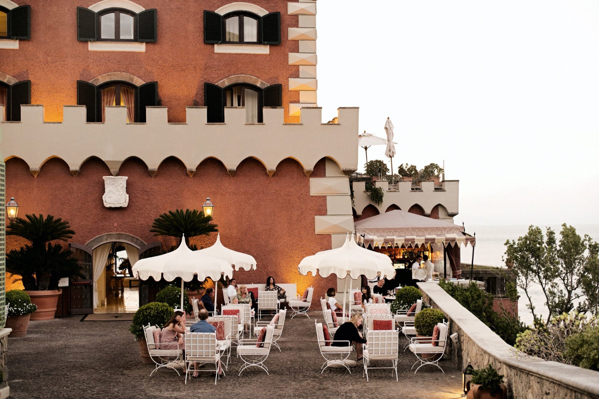 People sit at white tables with umbrellas on a terrace outside a large, orange-colored building with arched windows. The scene overlooks greenery and the sea under a bright sky. Some guests are dining or conversing. captured by seattle's best food and hospitality photographer Brooke Fitts
