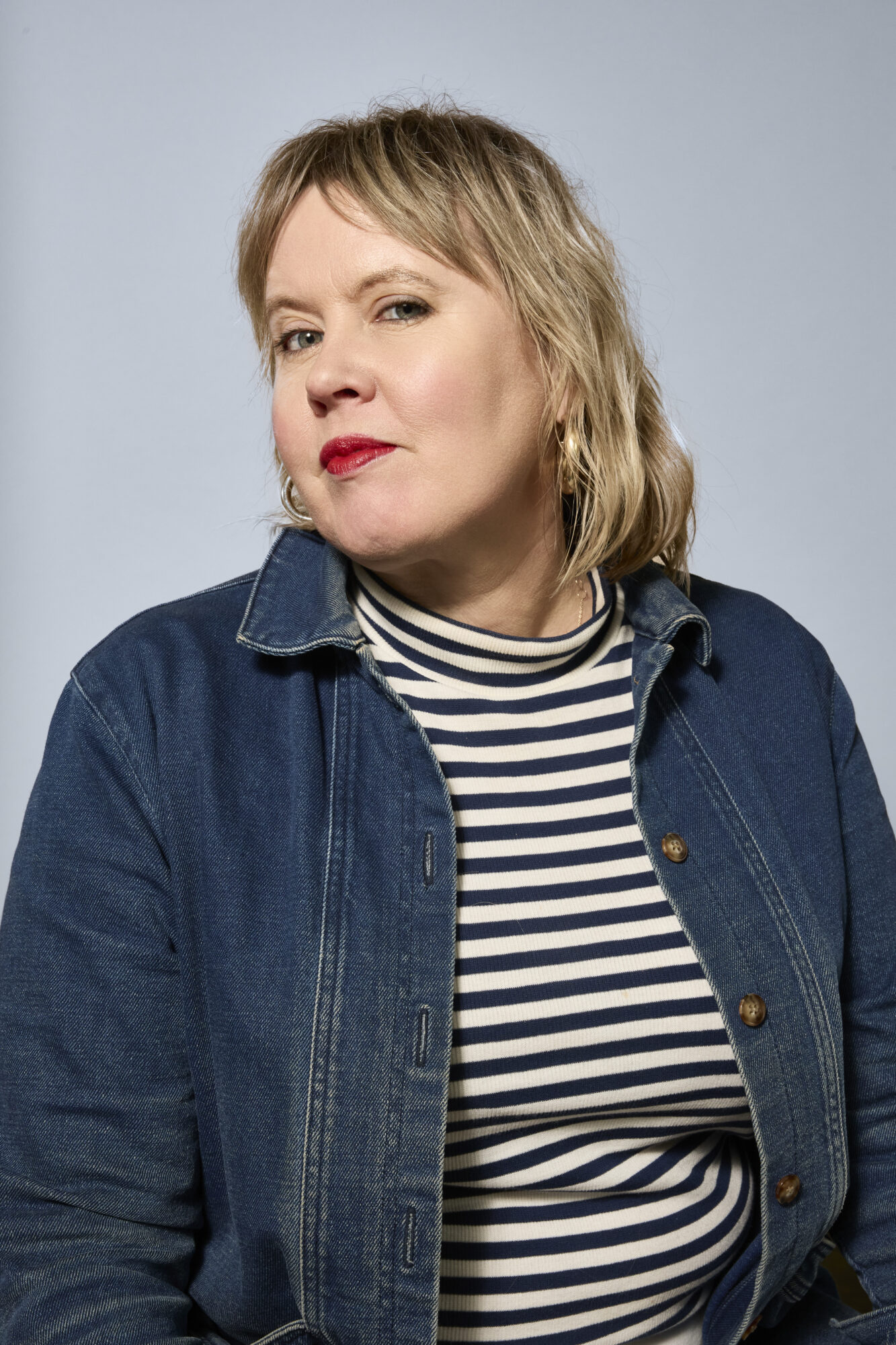 A person with blonde hair wearing red lipstick, a blue denim jacket, and a black-and-white striped shirt looks confidently at the camera against a plain light blue background, showcasing the eye for detail of a Seattle lifestyle photographer. captured by seattle's best food and hospitality photographer Brooke Fitts